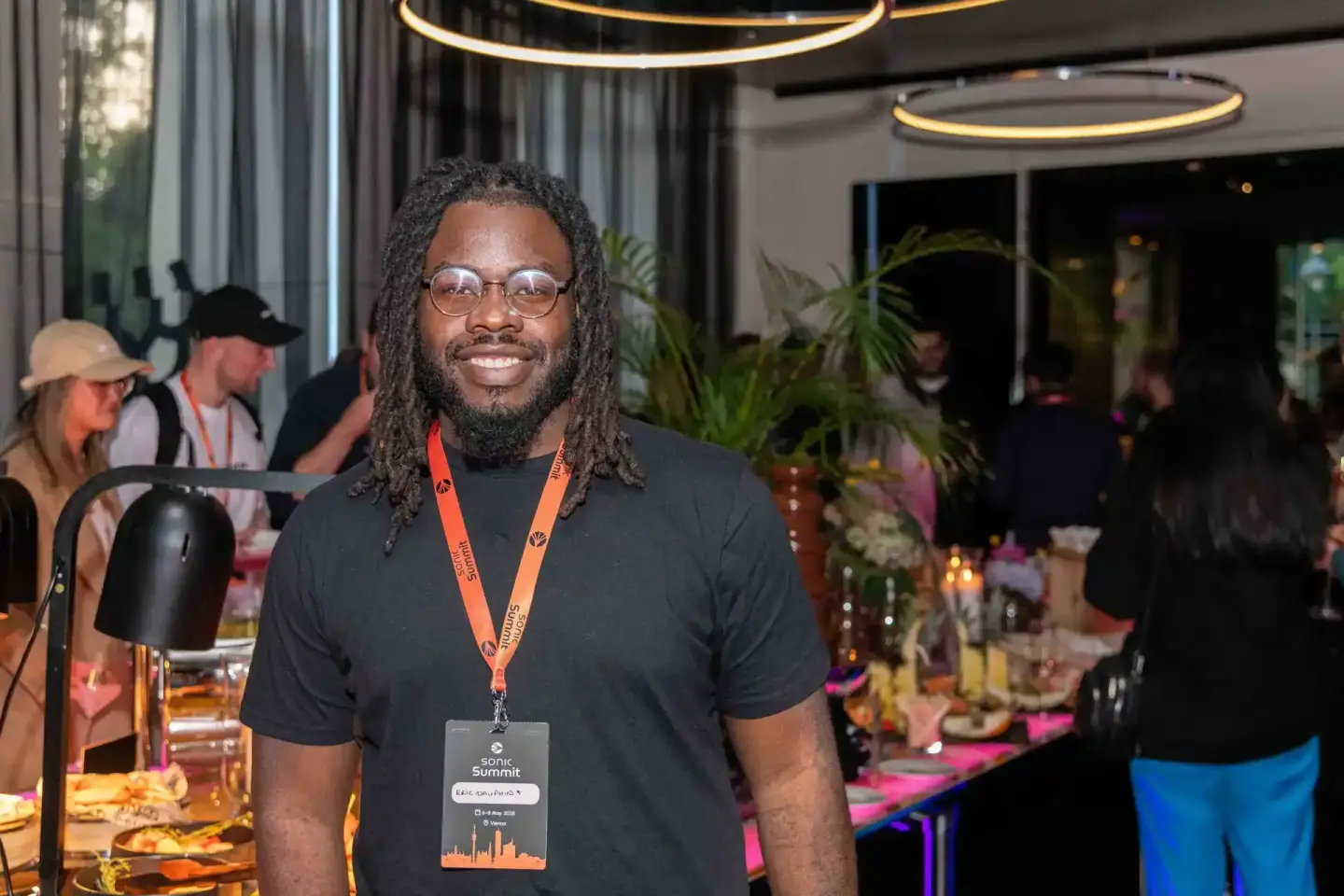 Lächelnder Mann mit Dreadlocks und Brille trägt schwarzes T-Shirt mit orange Konferenz-Badge bei Sonic Summit Eventfotografie