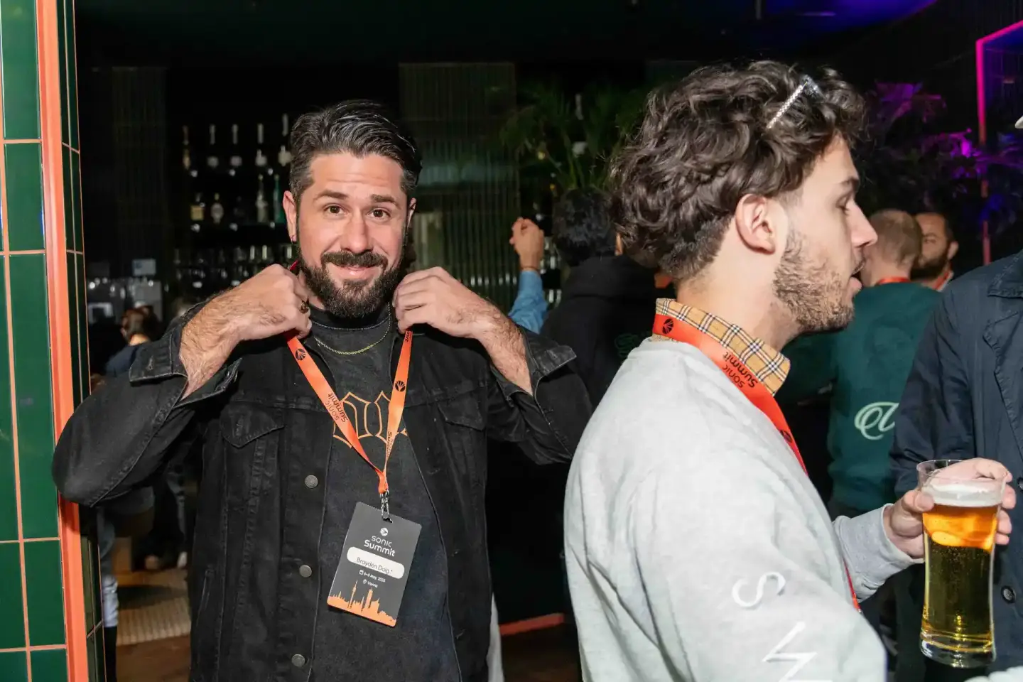 Zwei Männer beim Sonic Summit Event in Wien: Ein Mann mit schwarzer Jeansjacke und Eventpass lächelt in die Kamera, sein Gesp