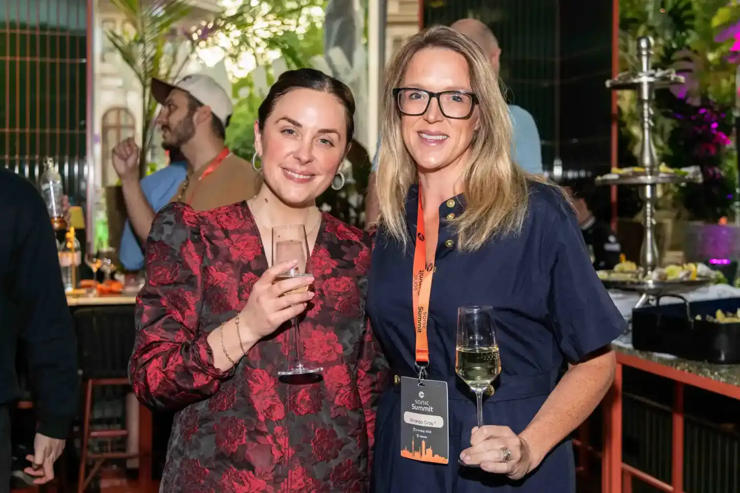 Zwei Frauen beim Sonic Summit Event in Wien, eine in rotem Muster-Outfit mit Getränk, eine in dunklem Hemd mit Eventpass und 