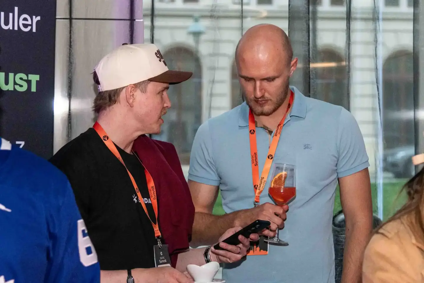Zwei Männer mit Ausweisen unterhalten sich auf dem Sonic Summit Event in Wien, einer hält ein Glas mit Getränk