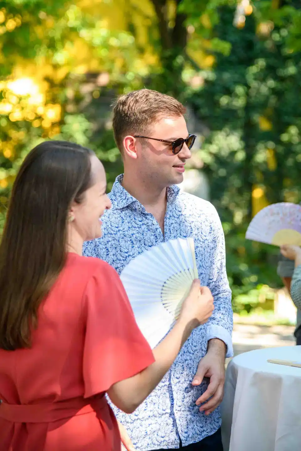 Mitarbeiterin im roten Kleid lacht ihren Kollegen an, beide mit Fächer beim Sommerfest im Wiener Gastgarten