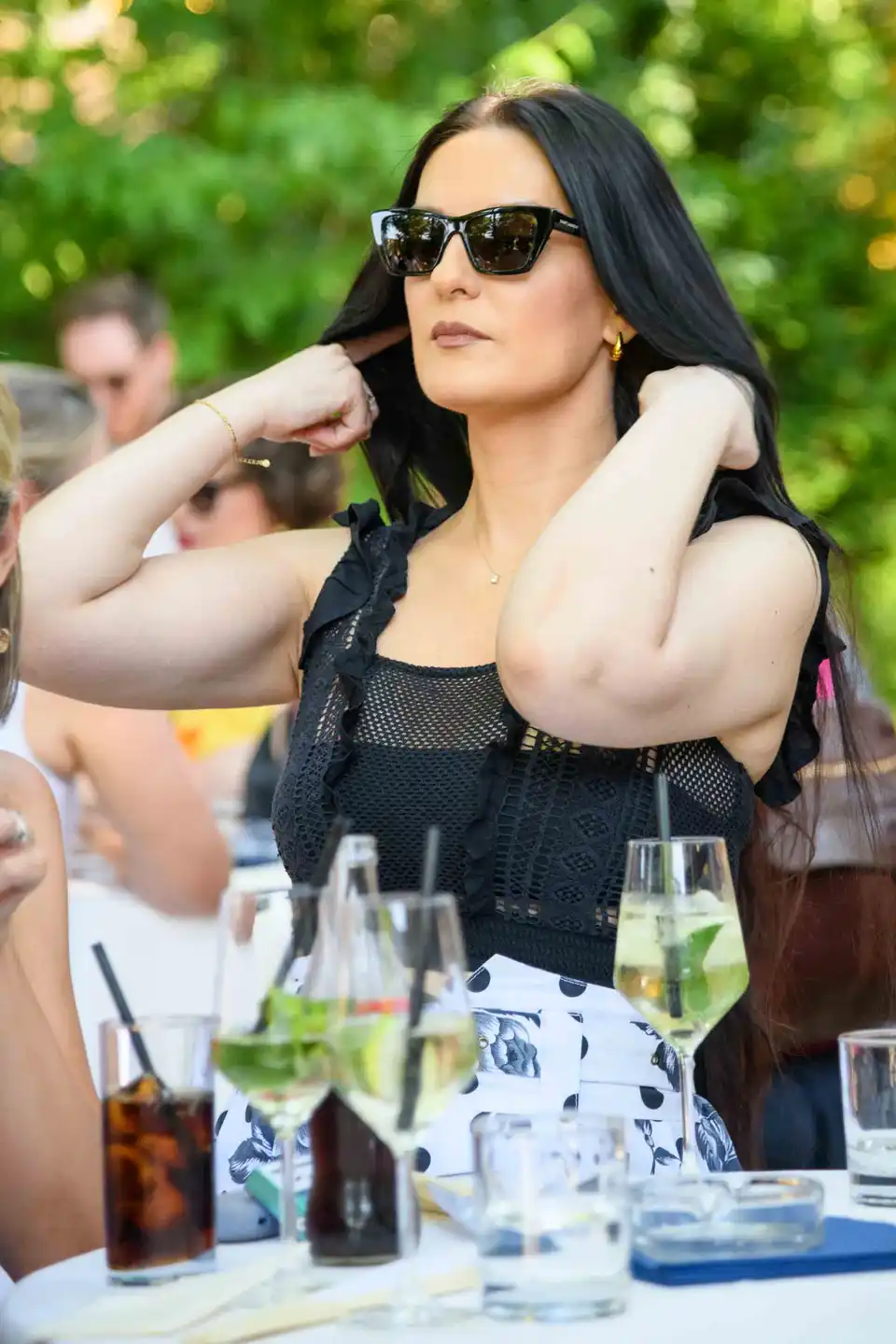 Gast mit Sonnenbrille und schwarzem Spitzentop richtet die Haare, davor Hugo-Gläser beim Sommerfest in Wien