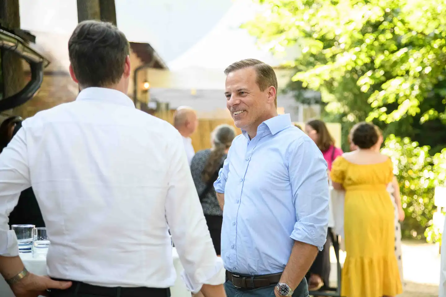 Mitarbeiter im hellblauen Hemd lächelt seinem Kollegen zu, dahinter Gäste am Stehtisch – Sommerfest Wien