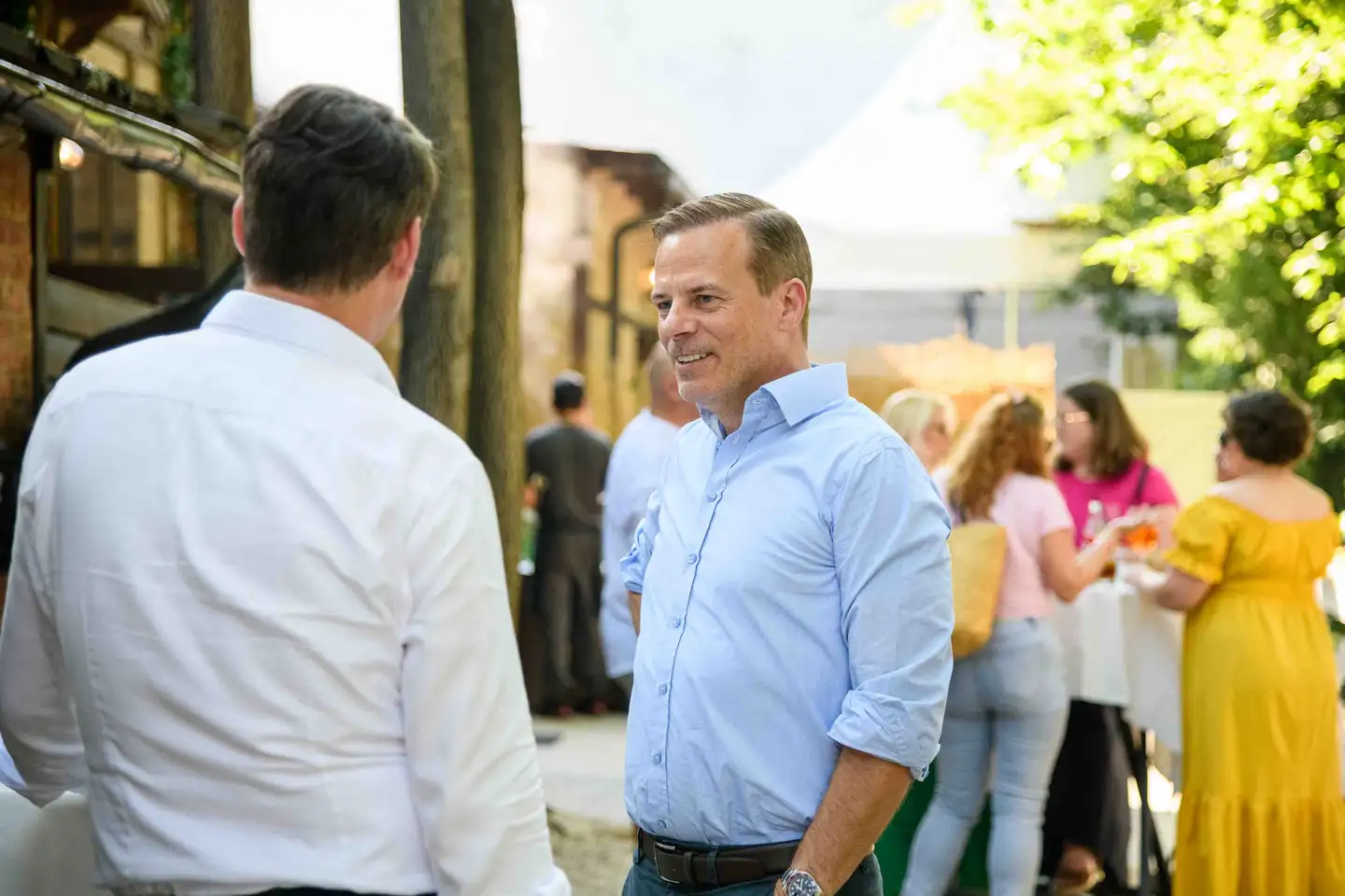Kollege im hellblauen Hemd im Gespräch, dahinter Gäste mit Aperol Spritz am Stehtisch – Firmenfeier Wien