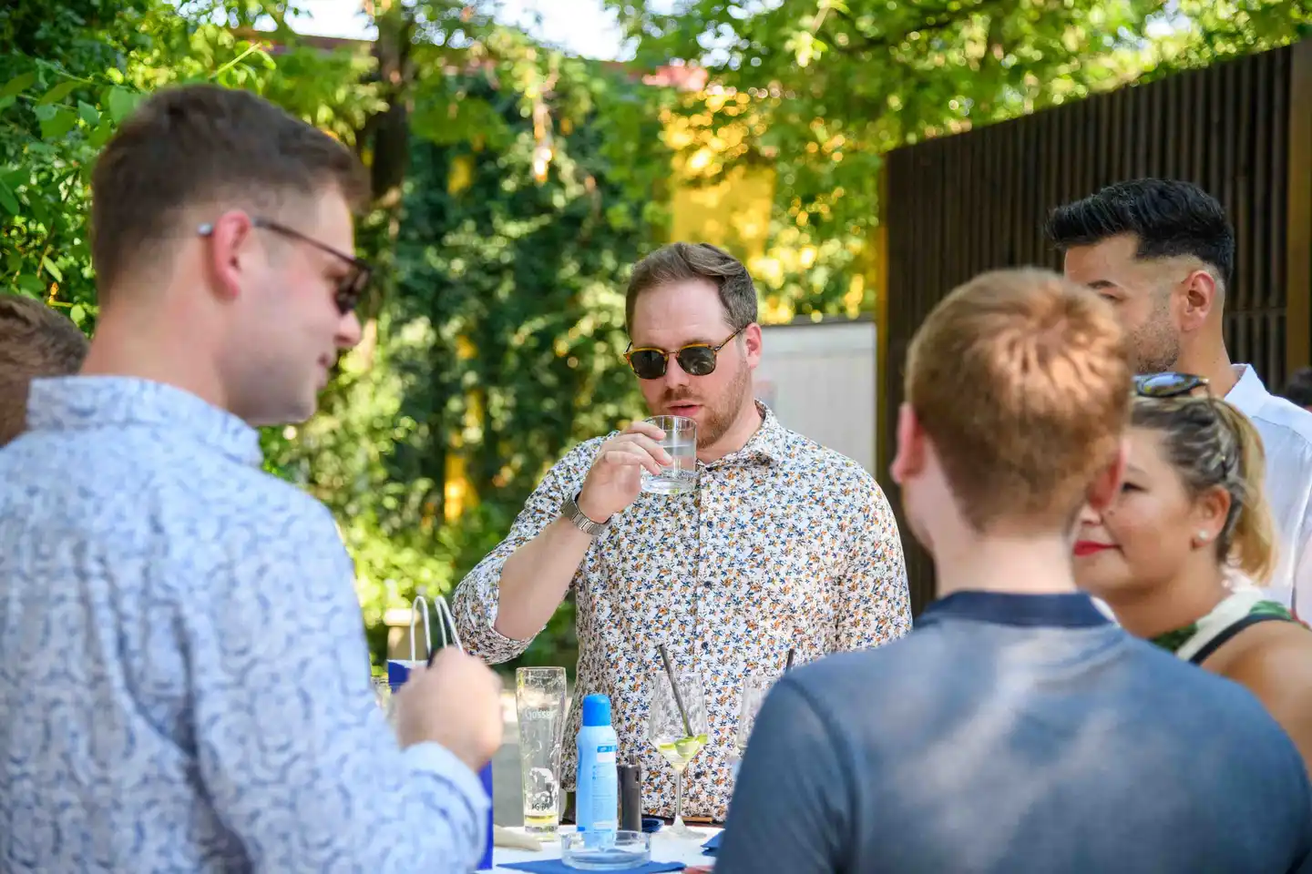 Mitarbeiter im geblümten Hemd trinkt Wasser am Stehtisch, umringt von Kollegen – Sommerfest Wien