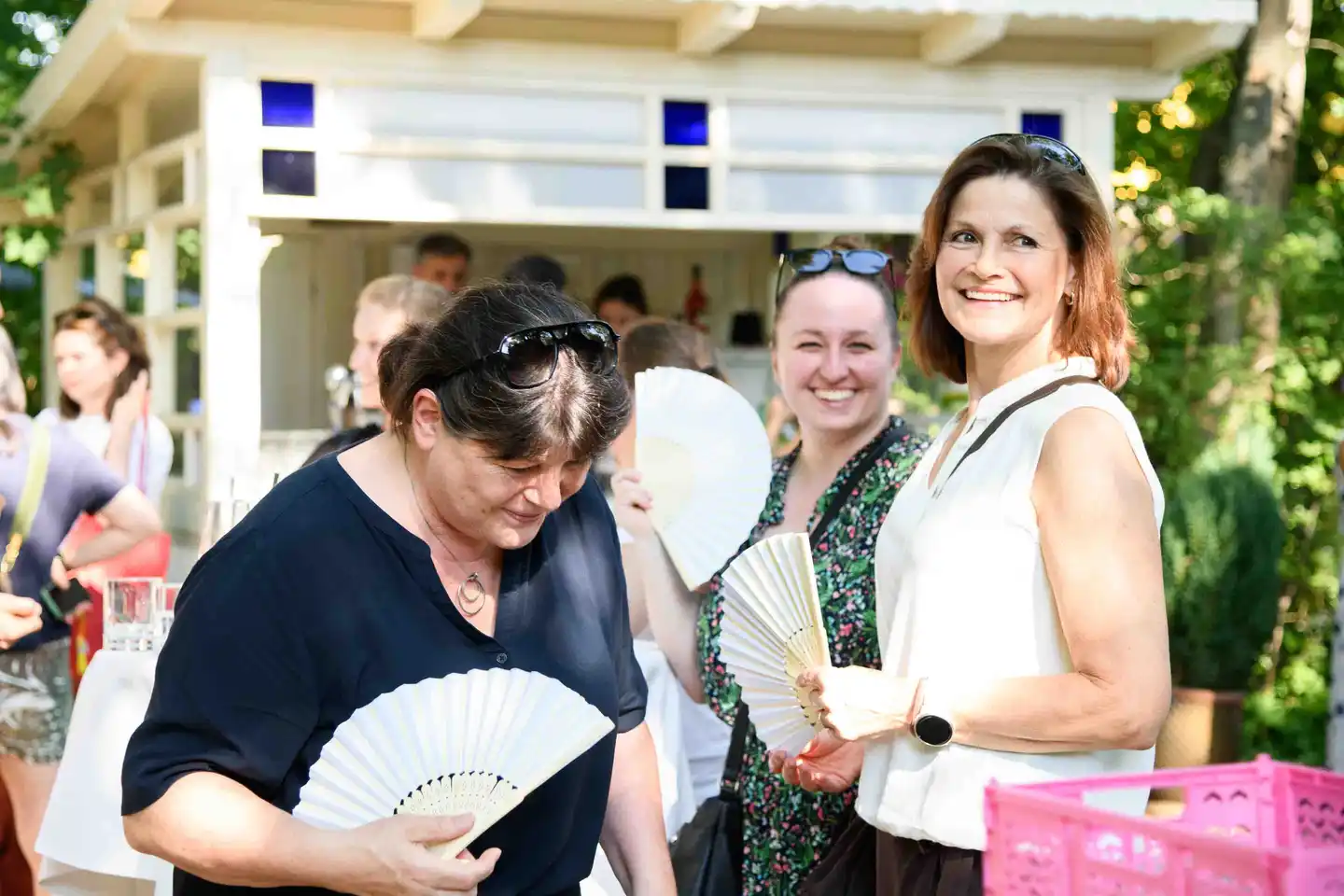 Drei Kolleginnen mit Papierfächern vor dem weißen Gartenpavillon beim Sommerfest in Wien