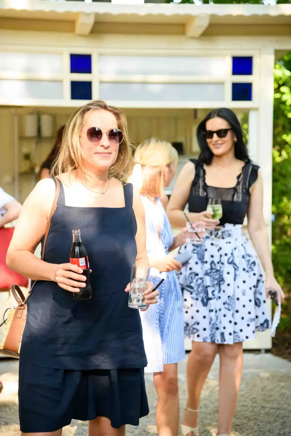 Kollegin im dunkelblauen Leinenkleid mit Cola-Flasche und Glas vor dem Gartenpavillon – Sommerfest Wien