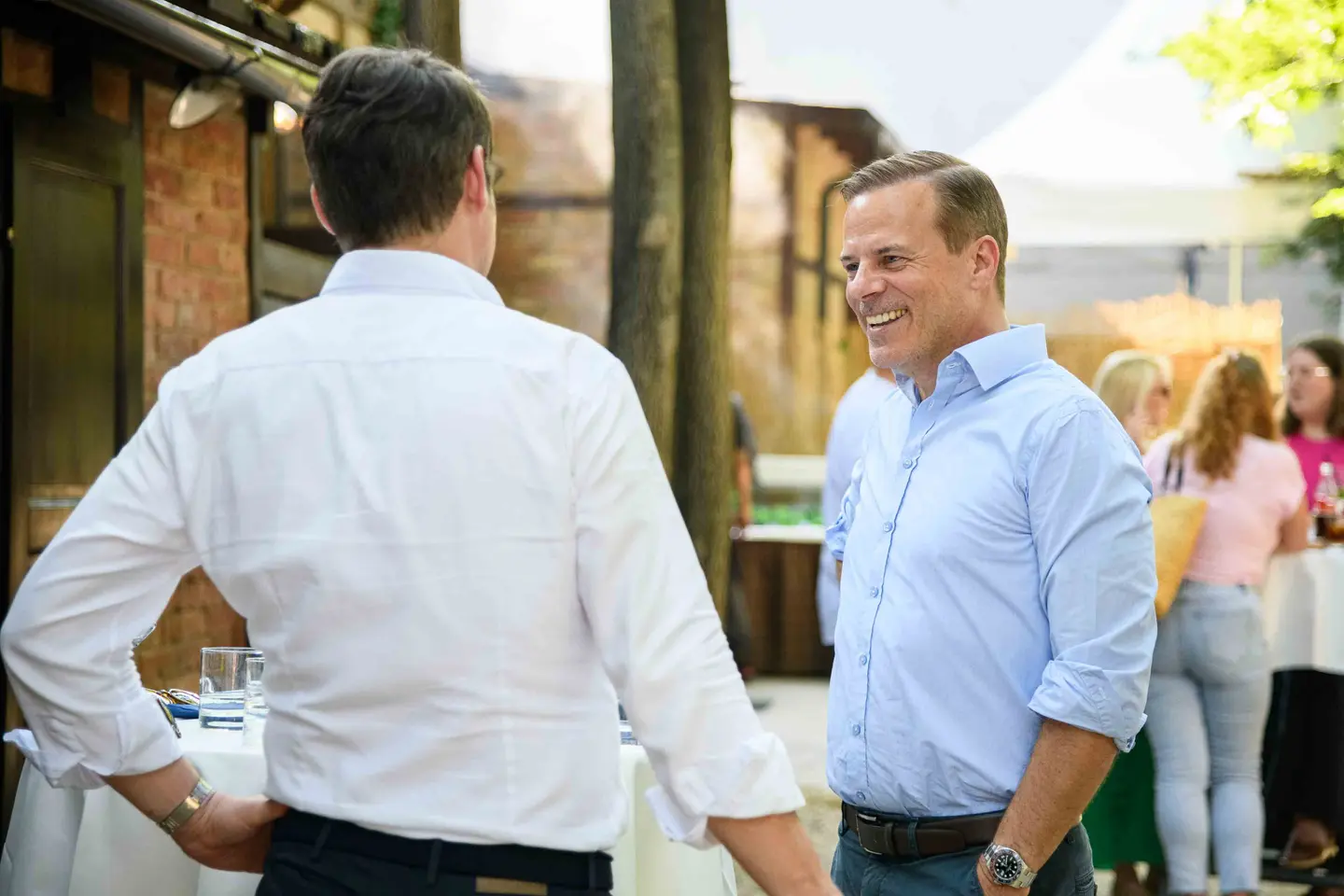 Kollege im blauen Hemd lacht im Gespräch vor Ziegelwand und Sonnensegel – Firmensommerfest Wien
