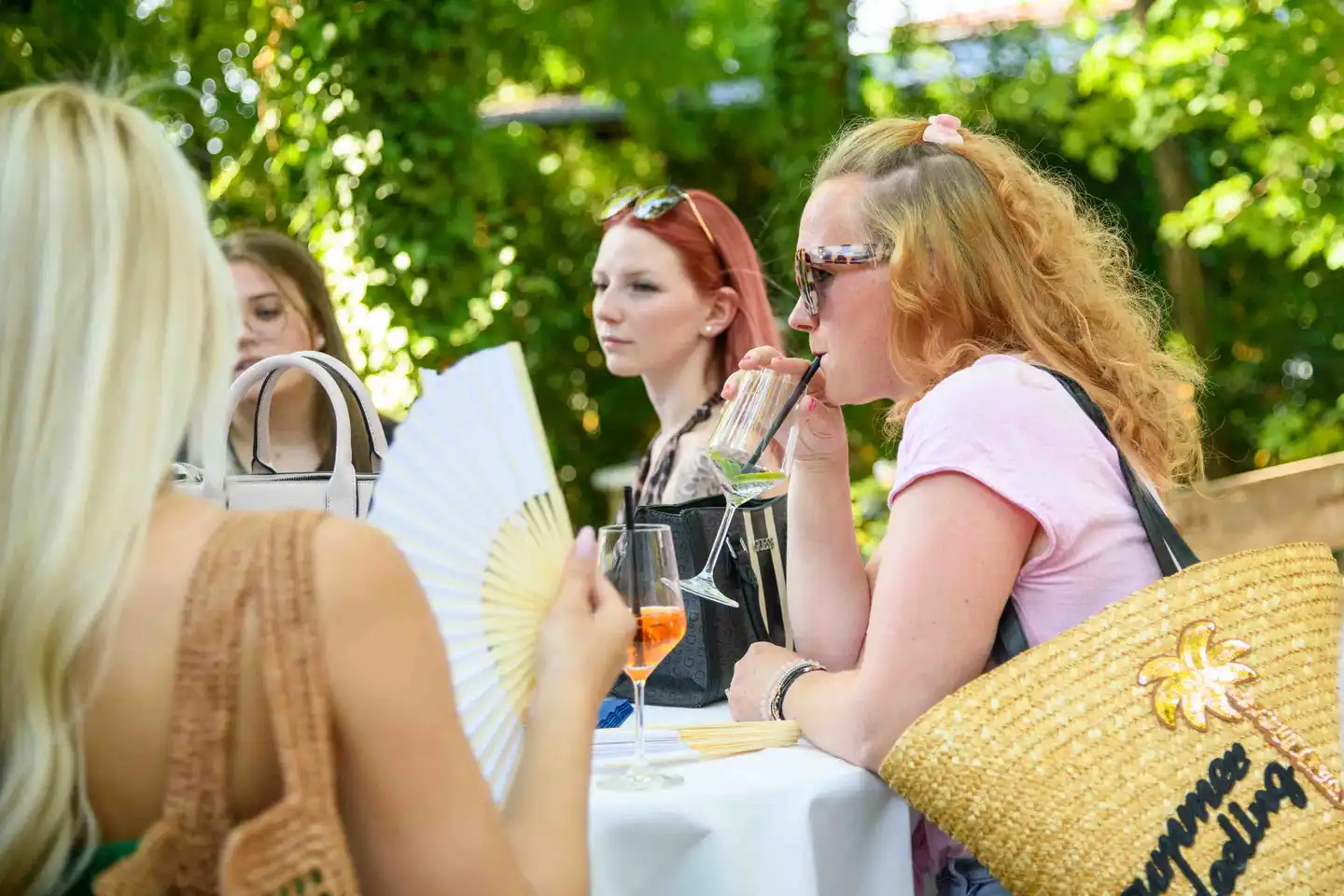 Kolleginnen am Stehtisch mit Fächer und Spritzer, eine trinkt mit Strohhalm – Sommerfest Wien