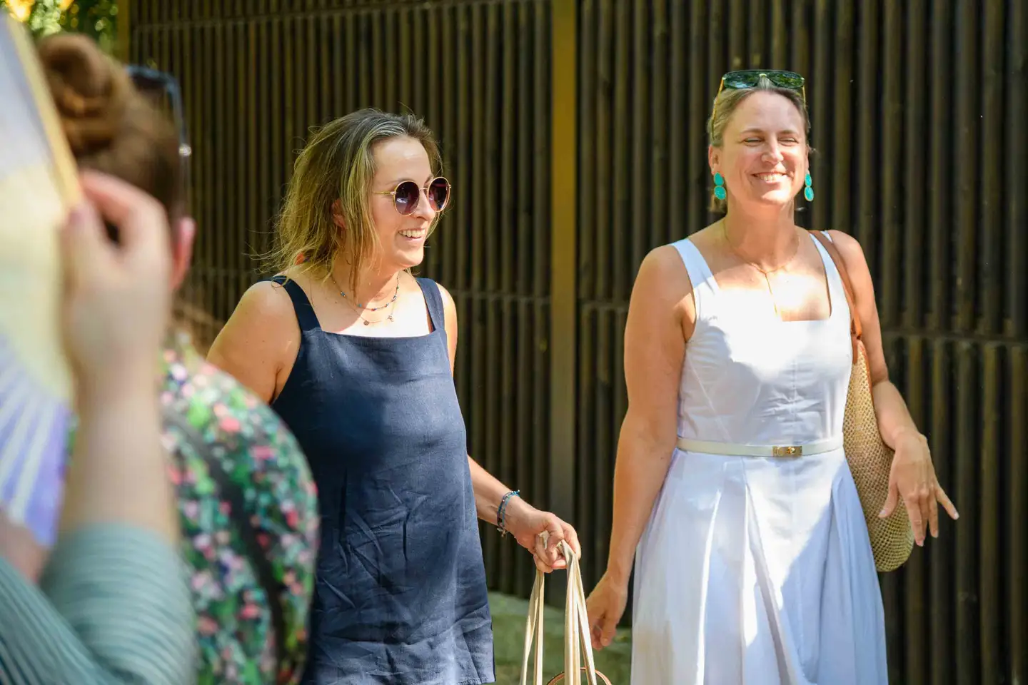 Zwei lachende Kolleginnen kommen mit Taschen zum Sommerfest, eine mit runder Sonnenbrille – Wien