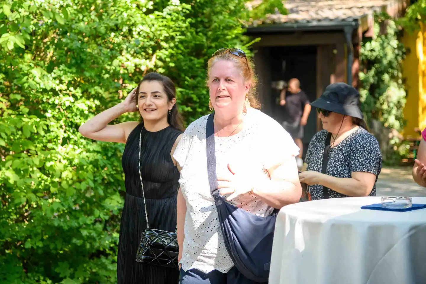 Zwei Gäste kommen in den Gastgarten, eine in weißer Lochstickerei-Bluse mit Umhängetasche – Sommerfest Wien