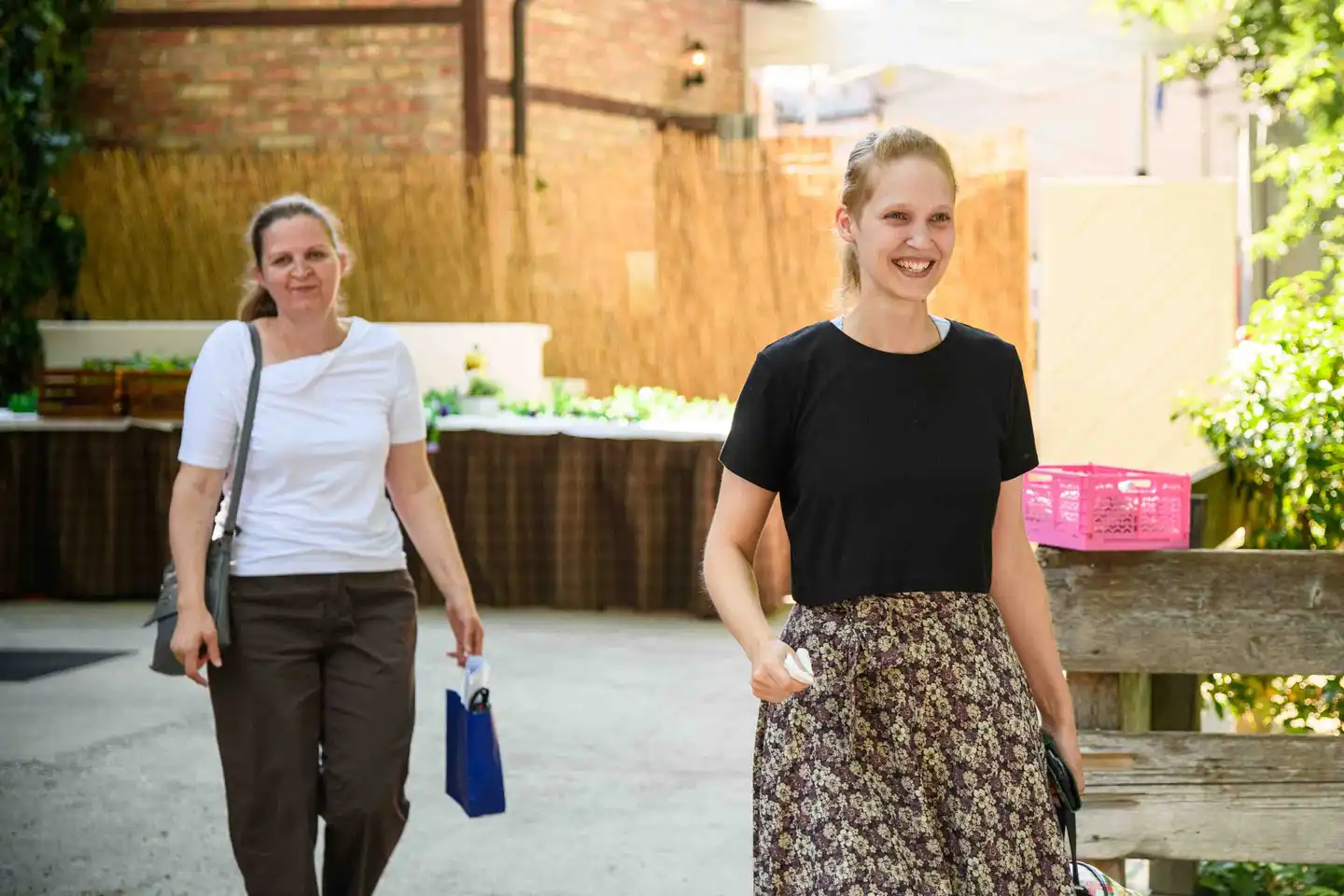 Zwei Mitarbeiterinnen kommen lachend über den Hof zum Sommerfest, eine mit blauem Geschenksackerl – Wien