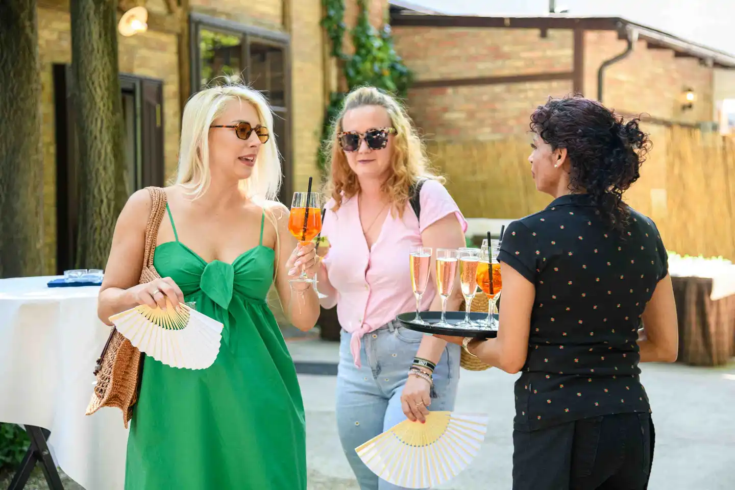 Servicekraft bringt Frizzante und Aperol zu zwei Gästen mit Fächern im Hof – Sommerfest Eventfotografie Wien