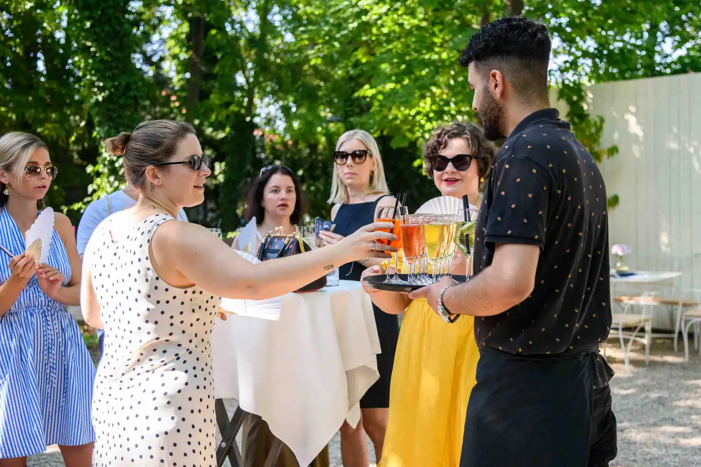 Mitarbeiterin im Punktekleid nimmt einen Aperol Spritz vom Tablett, Kolleginnen warten – Sommerfest Wien