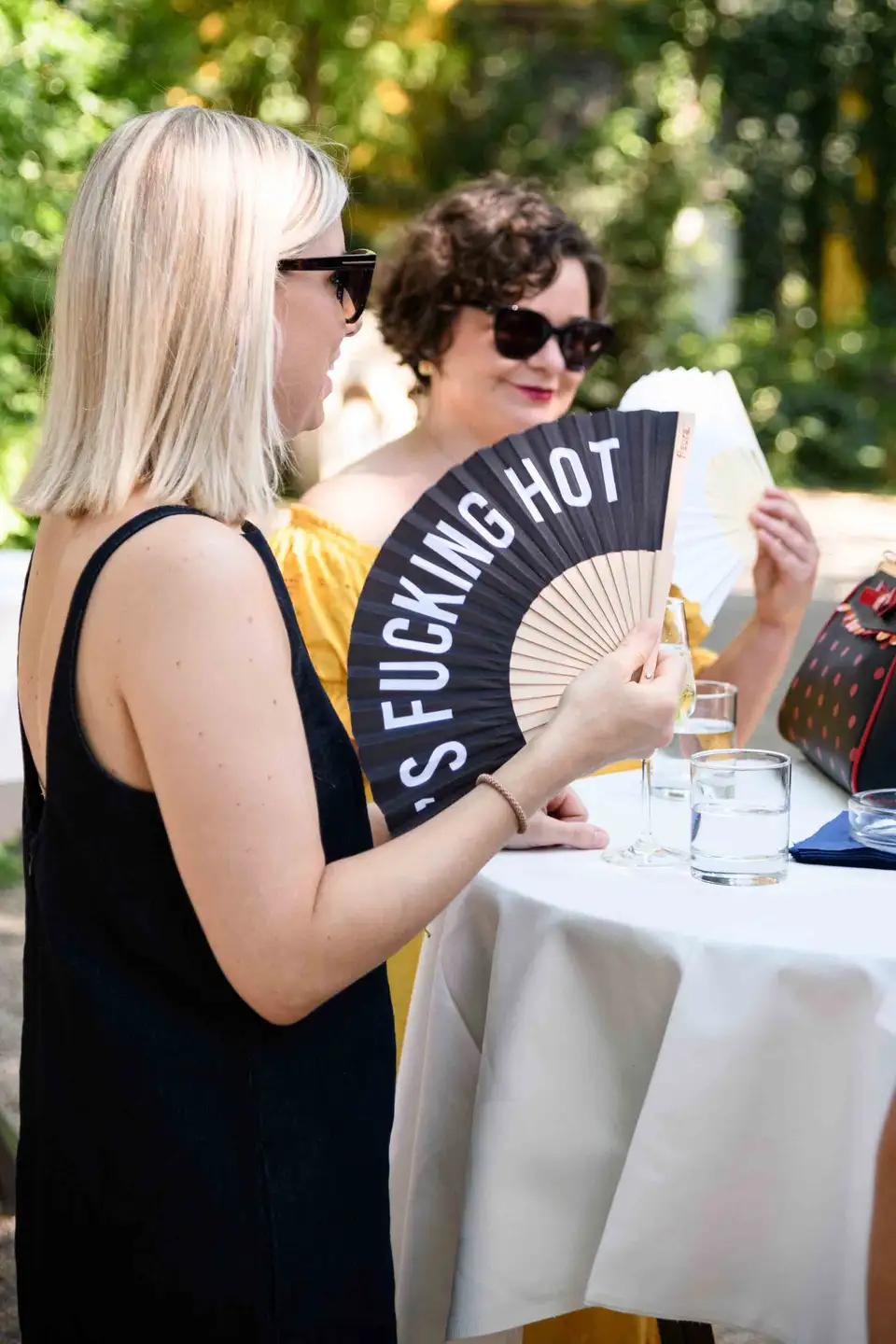 Gast im schwarzen Kleid hält einen bedruckten Fächer gegen die Hitze am Stehtisch – Sommerfest Wien