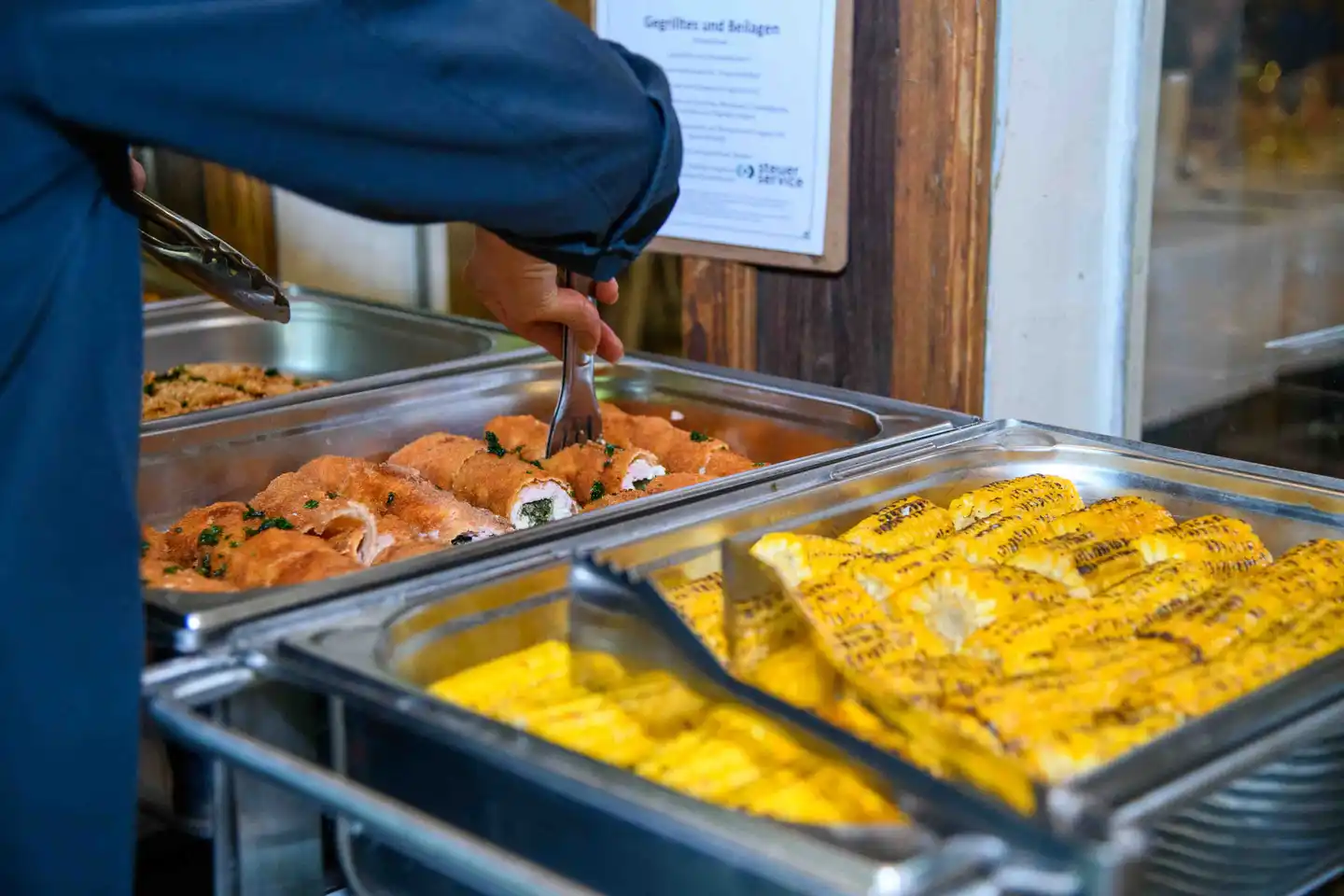 Servicemitarbeiter schöpft Cordon bleu am Buffet, daneben gegrillte Maiskolben – Sommerfest Wien