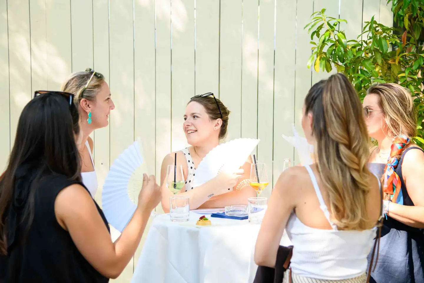 Vier Kolleginnen mit Fächern und Spritzer am Stehtisch vor heller Holzwand – Sommerfest Eventfotografie Wien