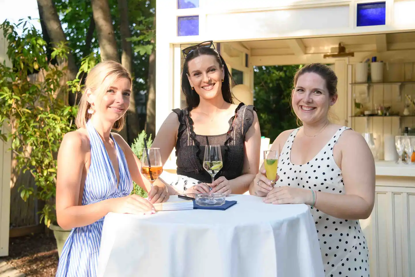 Drei lächelnde Kolleginnen am Stehtisch mit Aperol, Hugo und Fruchtcocktail – Sommerfest Wien