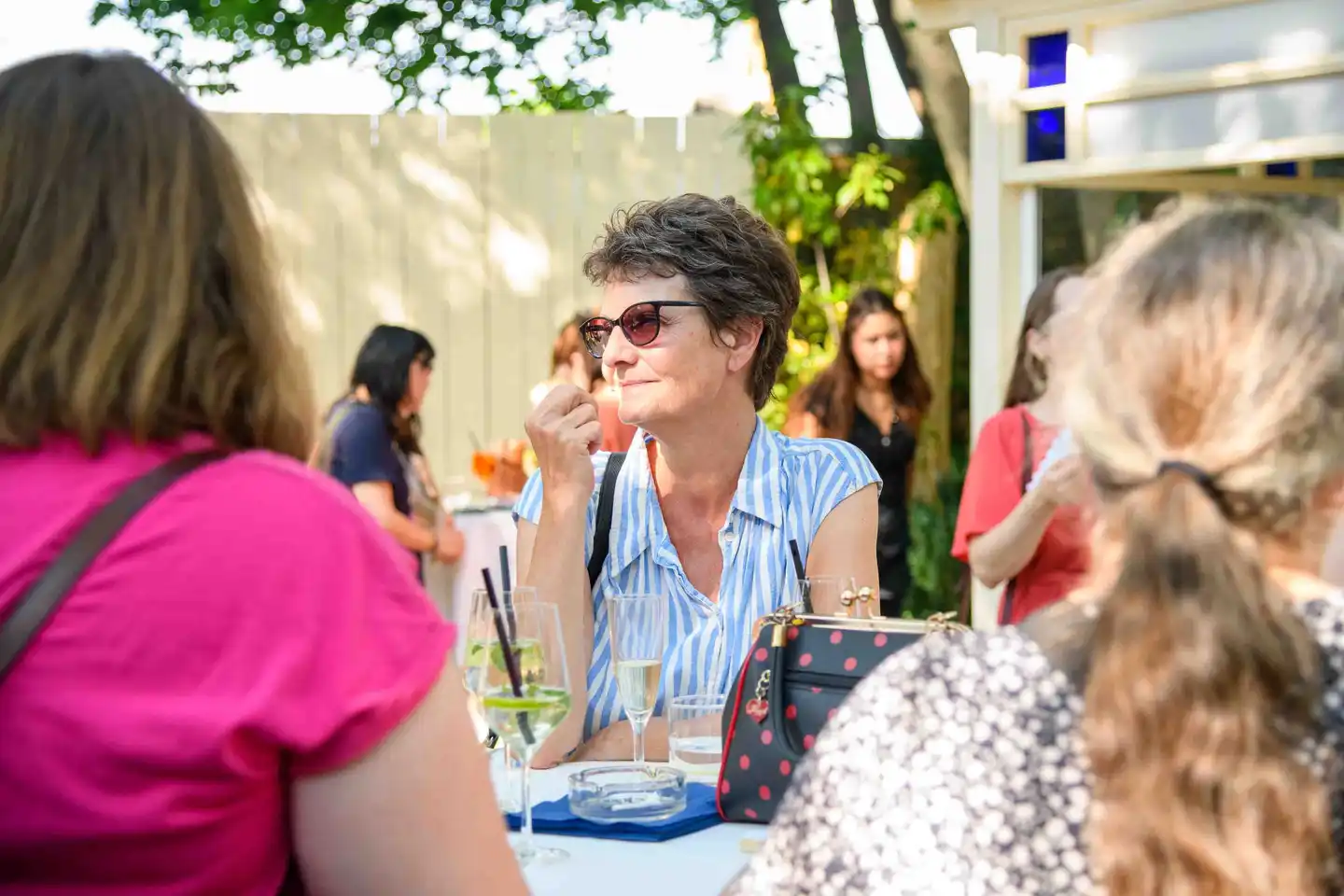 Gast im blau gestreiften Hemd mit Sonnenbrille am Stehtisch, Frizzante und Handtasche daneben – Sommerfest Wien