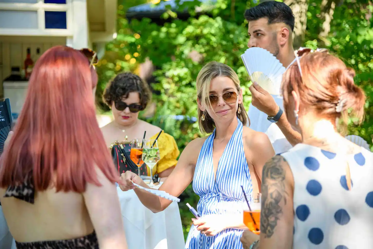 Gast im blau gestreiften Sommerkleid mit Fächer und Spritzer inmitten der Gästegruppe – Sommerfest Wien