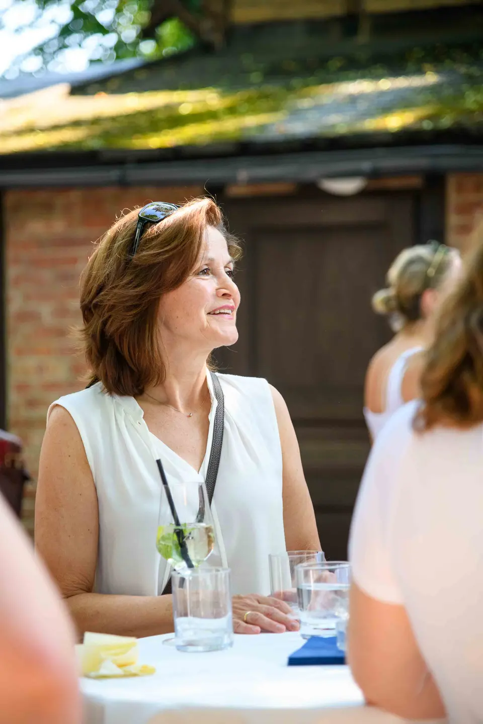 Gast in weißer Bluse im Profil, Sonnenbrille im Haar, am Stehtisch vor der Ziegelwand – Sommerfest Wien