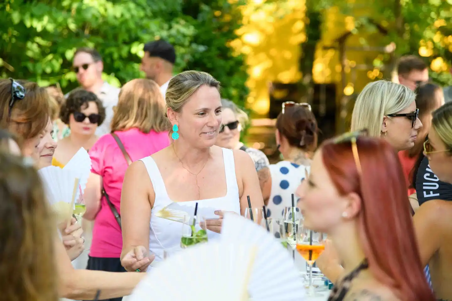 Gast im weißen Top mit türkisen Ohrringen inmitten der Gästemenge mit Fächern – Sommerfest Wien
