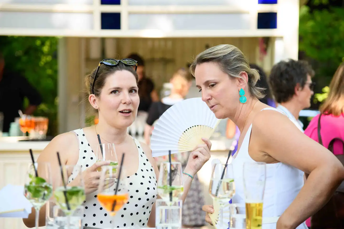 Zwei Kolleginnen im Gespräch an der Gartenbar, eine mit Fächer, Getränke stehen bereit – Sommerfest Wien