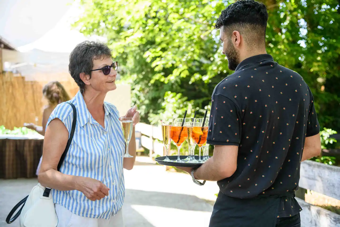 Gast im blau gestreiften Hemd im Gespräch mit dem Kellner samt Aperitif-Tablett – Sommerfest Wien