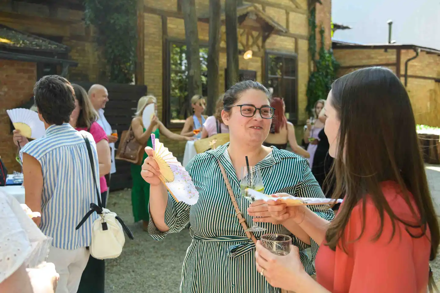 Mitarbeiterin im grün gestreiften Kleid zeigt ihren bemalten Fächer, dahinter der volle Gastgarten – Wien