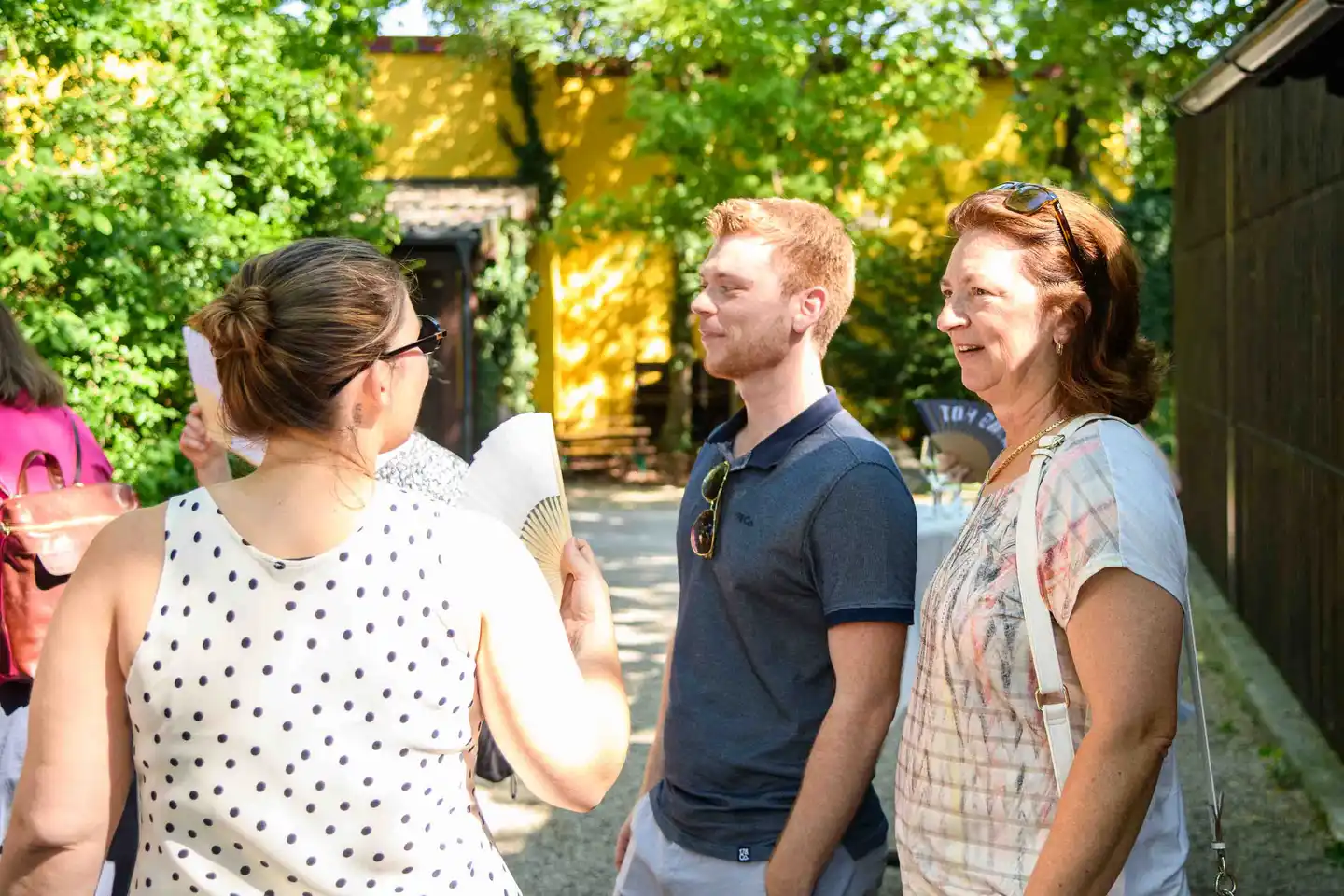 Drei Gäste im Gespräch am Kiesweg vor gelber Hauswand, eine Frau mit weißem Fächer – Sommerfest Wien