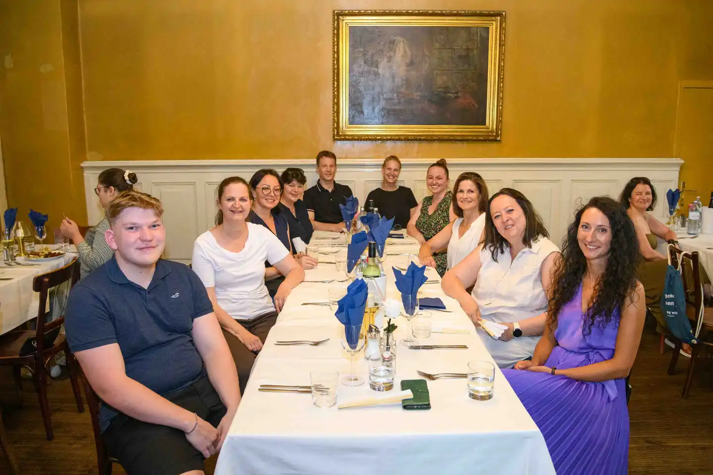 Team an der langen Tafel im Restaurantsaal mit blauen Servietten lächelt in die Kamera – Sommerfest Wien