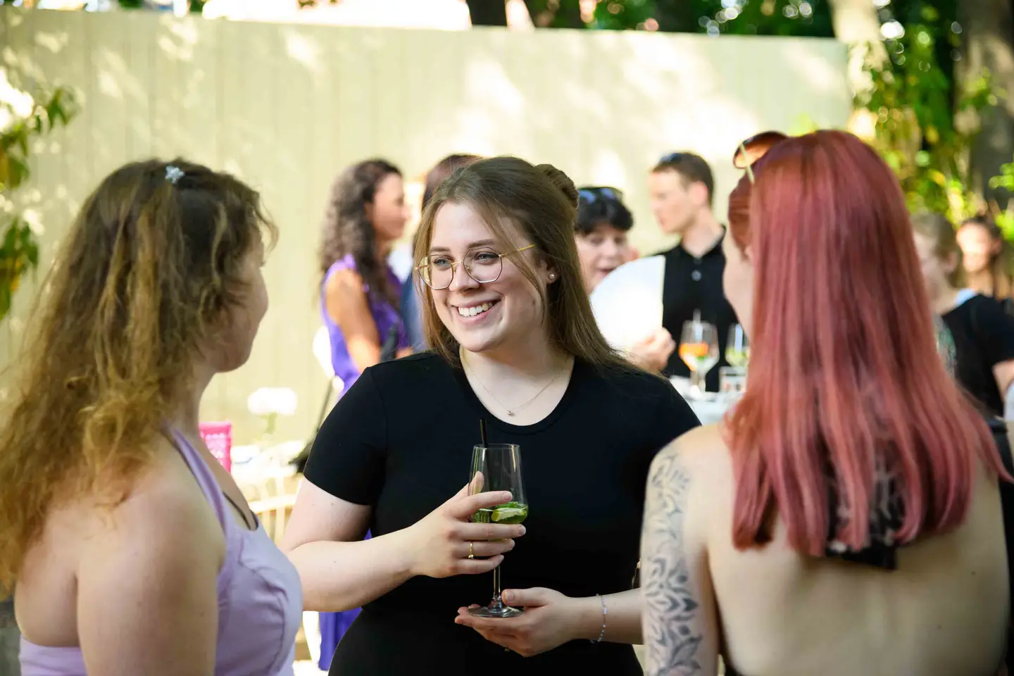 Junge Mitarbeiterin mit Brille hält einen Weißweinspritzer und lacht im Gespräch – Sommerfest Eventfotografie Wien