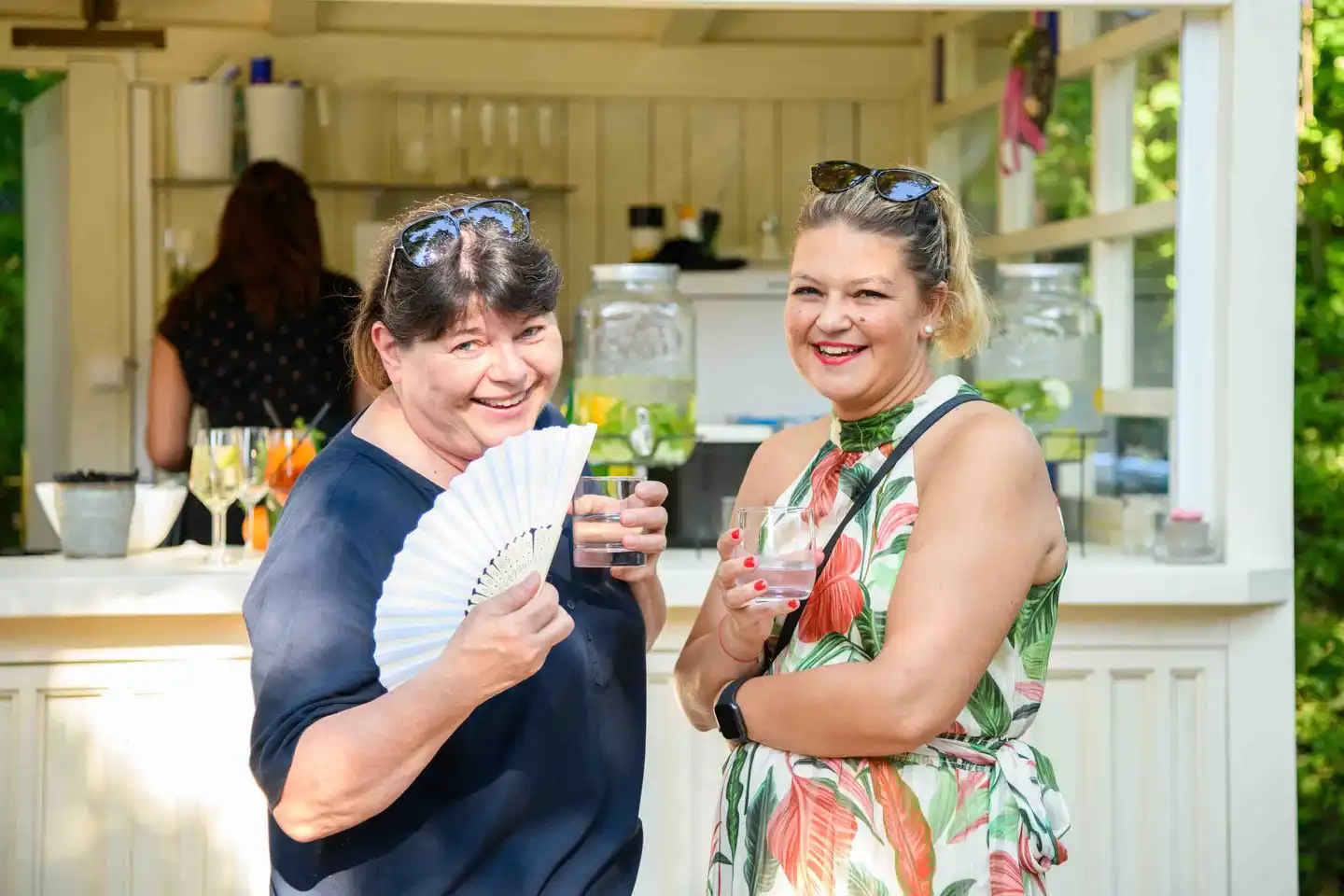 Zwei Kolleginnen mit Wassergläsern und Fächer posieren vor der weißen Gartenbar – Sommerfest Wien