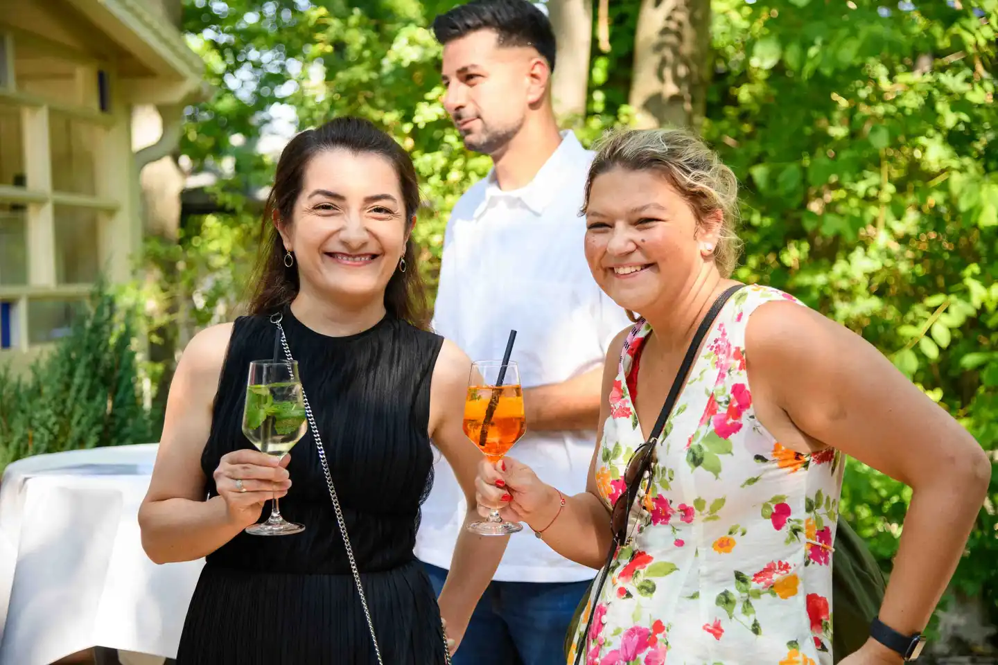 Zwei Kolleginnen prosten mit Hugo und Aperol Spritz in die Kamera, Kollege im Hintergrund – Sommerfest Wien
