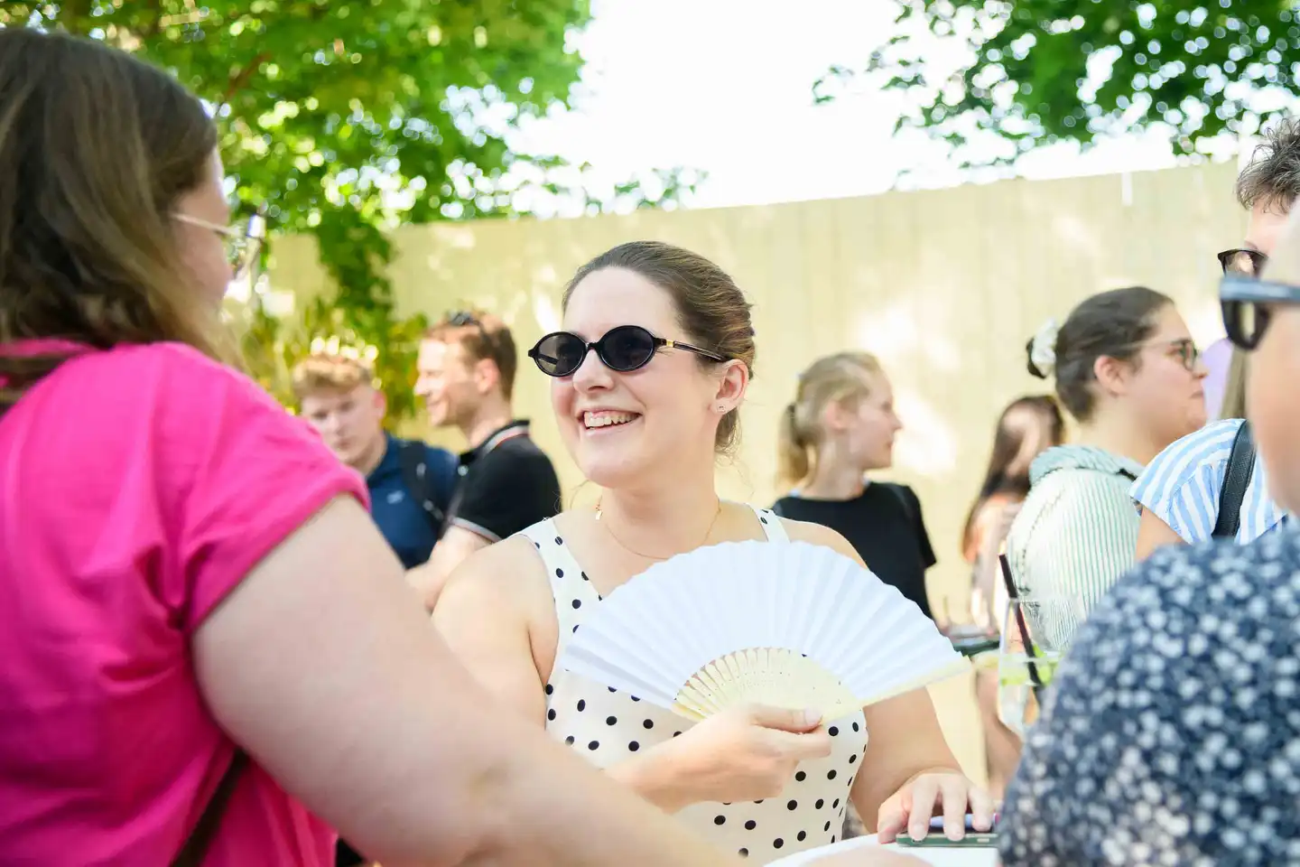 Lachende Kollegin mit Sonnenbrille und weißem Fächer im Punktekleid – Sommerfest Eventfotografie Wien