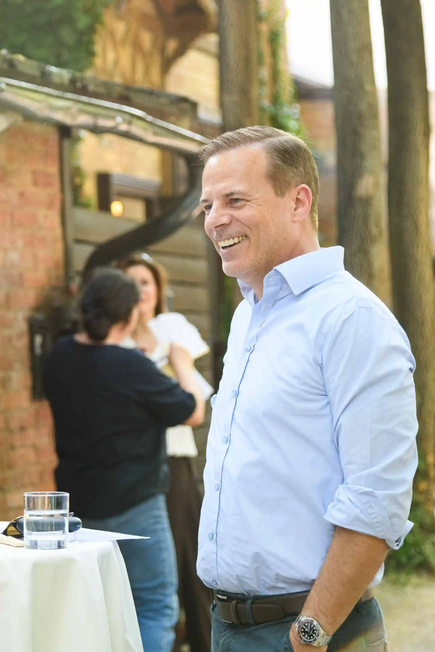 Lachender Gast im hellblauen Hemd am Stehtisch vor der Ziegelfassade – Sommerfest Eventfotografie Wien