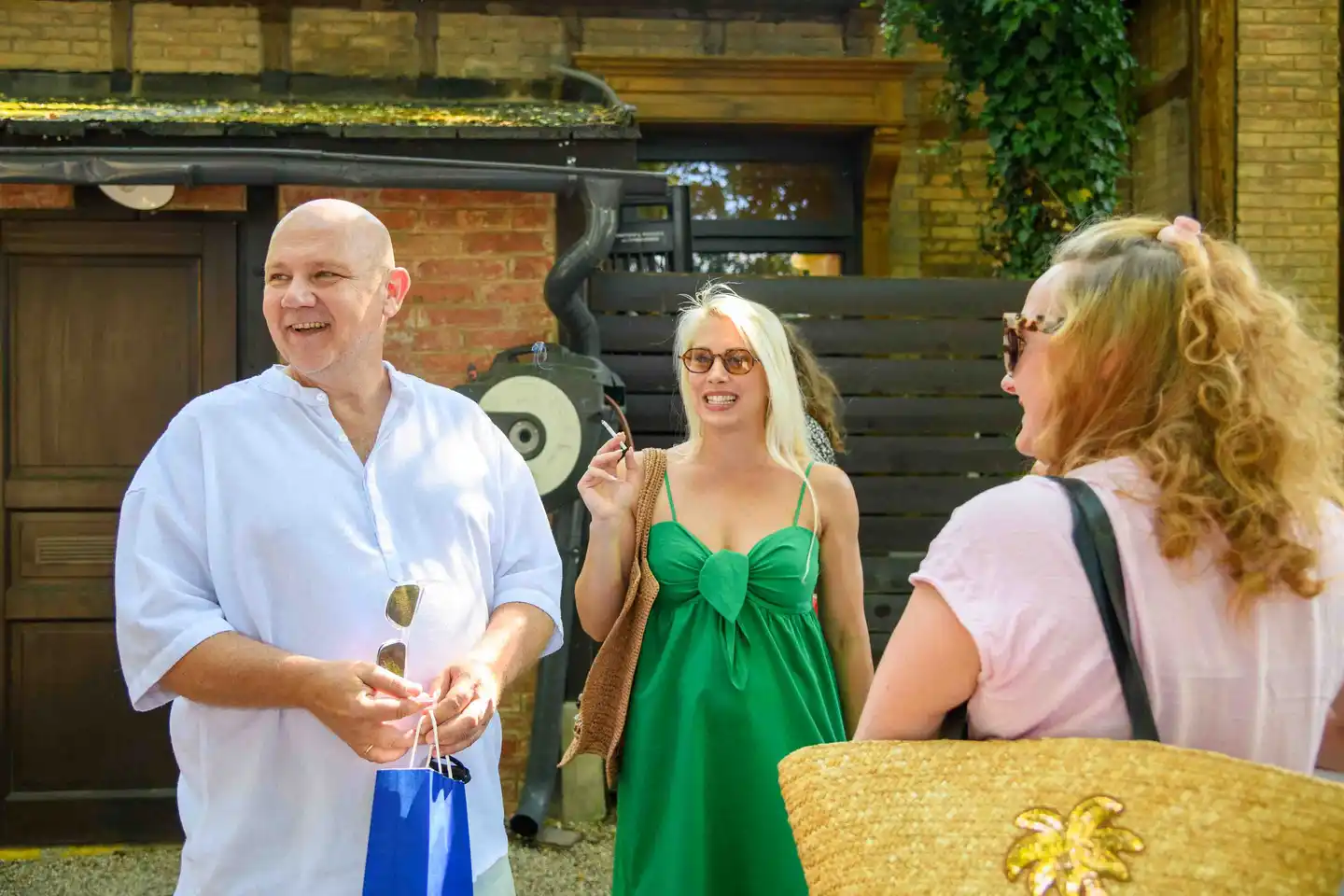 Lachender Gast mit blauem Geschenksackerl im Gespräch mit zwei Kolleginnen vor Ziegelwand – Sommerfest Wien