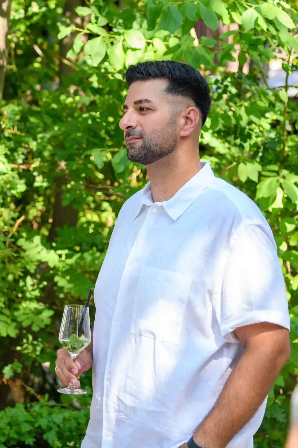 Mitarbeiter im weißen Leinenhemd mit Hugo-Glas vor grüner Hecke beim Sommerfest in Wien