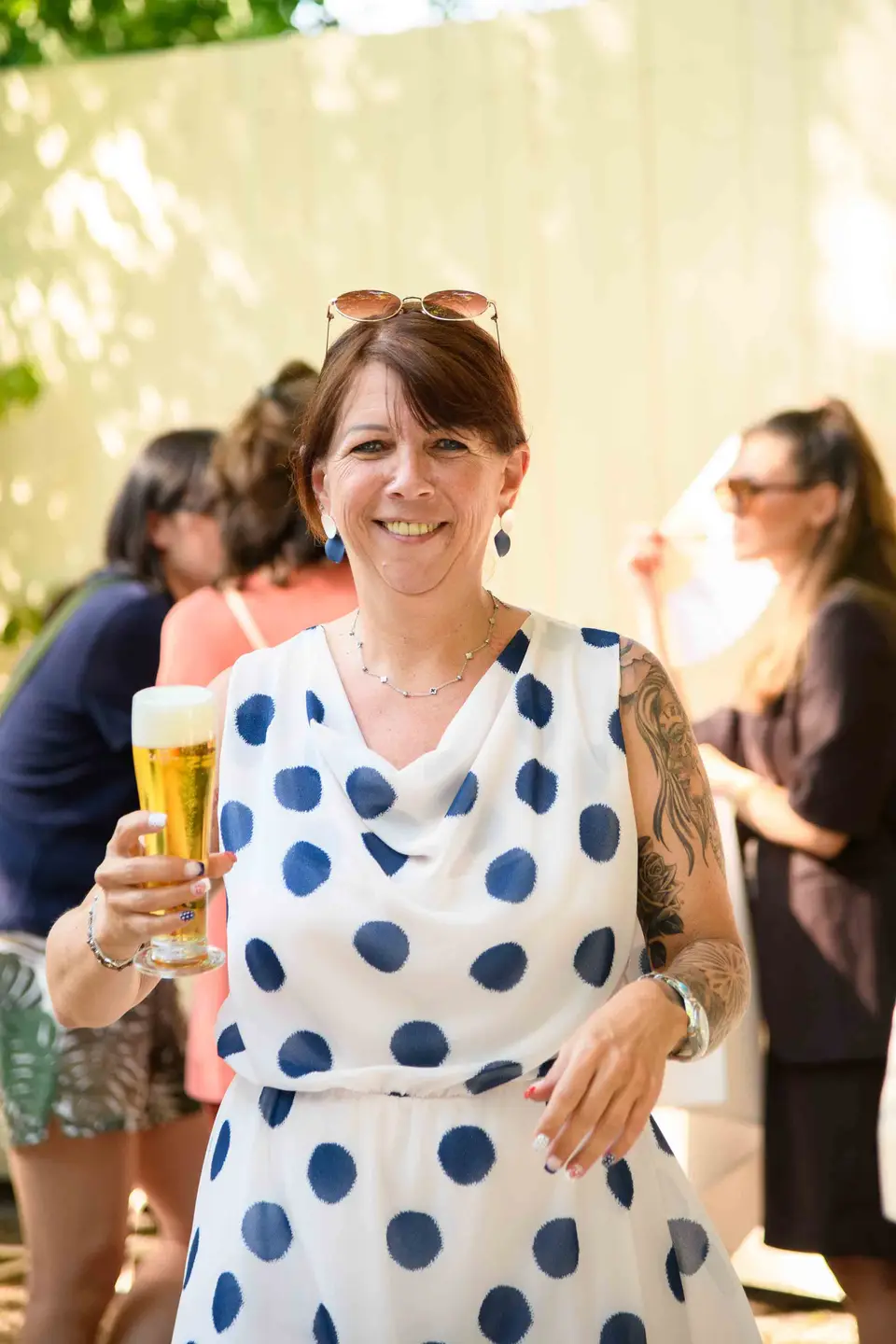 Mitarbeiterin im weiß-blauen Punktekleid hält ein Glas Bier und lächelt in die Kamera – Sommerfest Wien