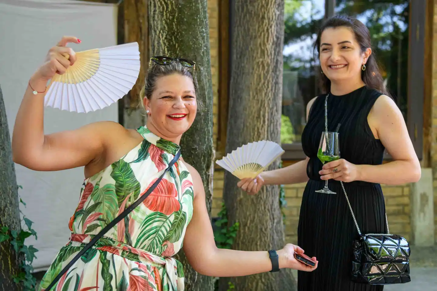 Zwei Kolleginnen posieren lachend mit weißen Fächern unter den Bäumen im Hof – Sommerfest Wien