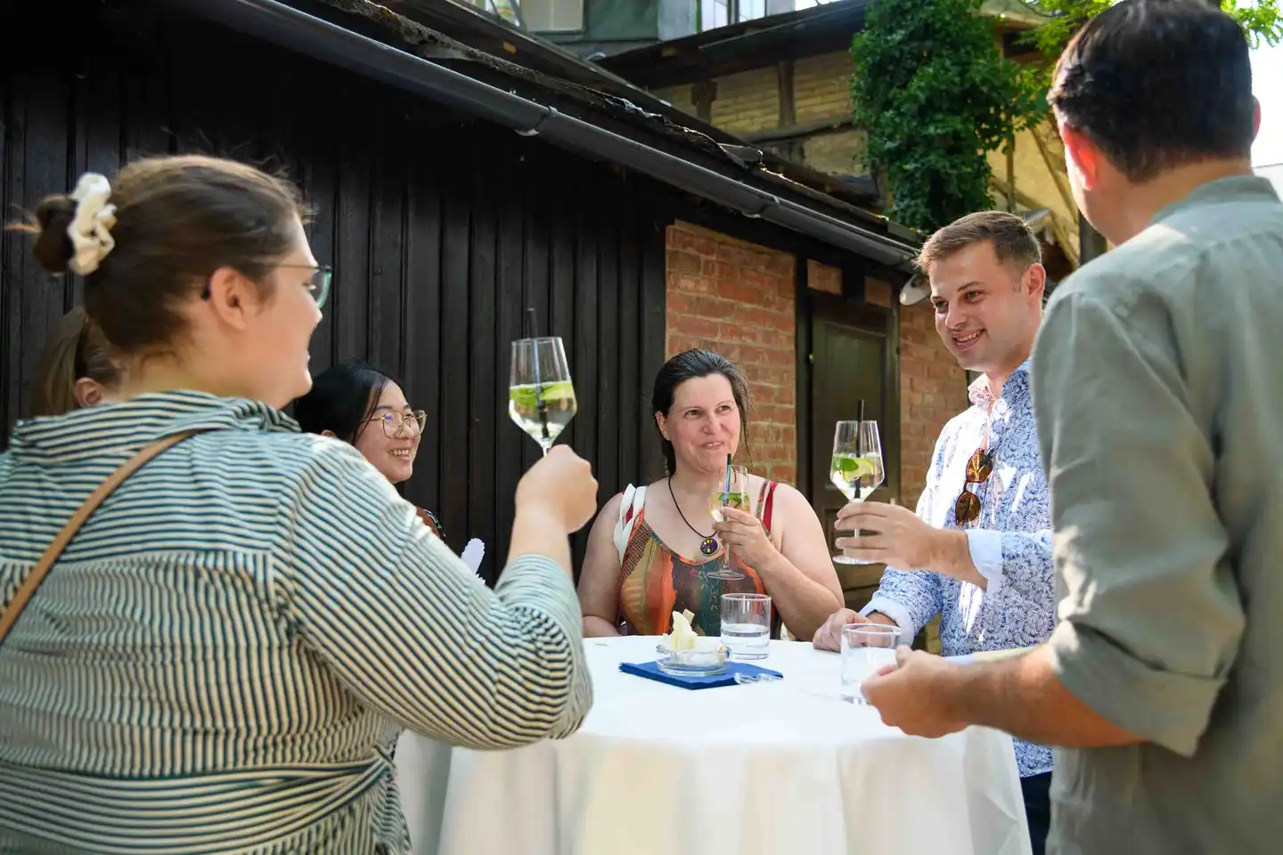 Gemischte Runde am Stehtisch hebt Hugo-Gläser vor der schwarzen Holzwand – Sommerfest Eventfotografie Wien