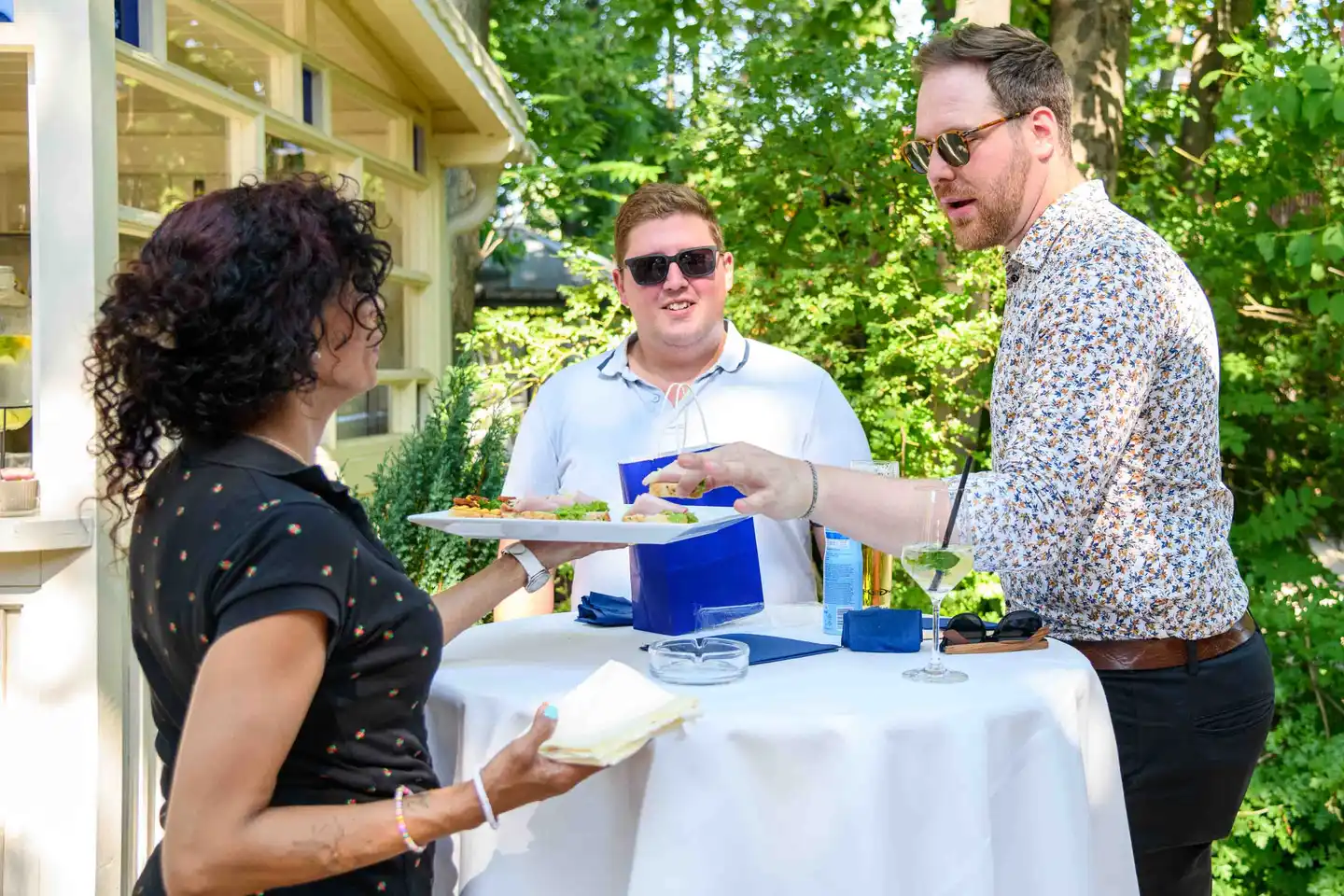 Servicekraft reicht zwei Gästen eine Platte mit Fingerfood am Stehtisch – Sommerfest Eventfotografie Wien
