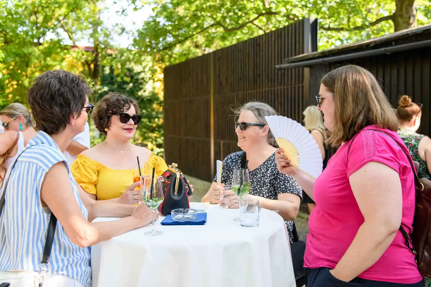 Vier Kolleginnen mit Spritzer am Stehtisch im Gastgarten, eine kühlt sich mit Fächer – Sommerfest Eventfotografie Wien