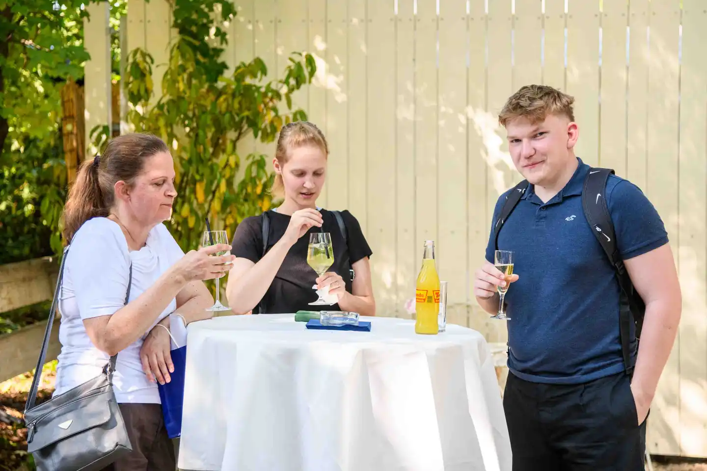 Drei Kollegen am Stehtisch mit Weißweinspritzer und Frizzante vor hellem Holzzaun – Sommerfest Wien