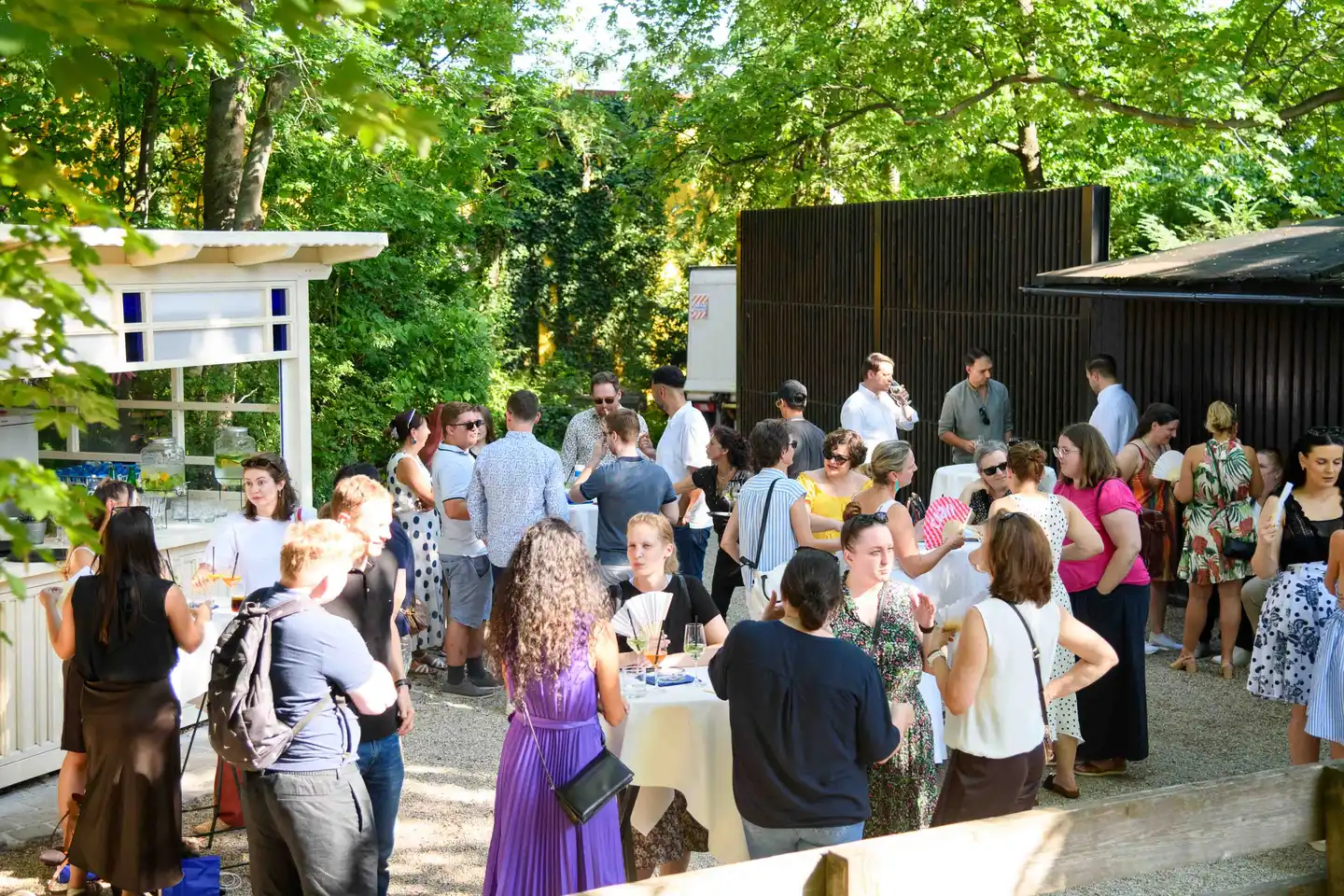 Überblick über das Firmensommerfest: Gäste an Stehtischen im Gastgarten mit Bar im weißen Pavillon, Wien