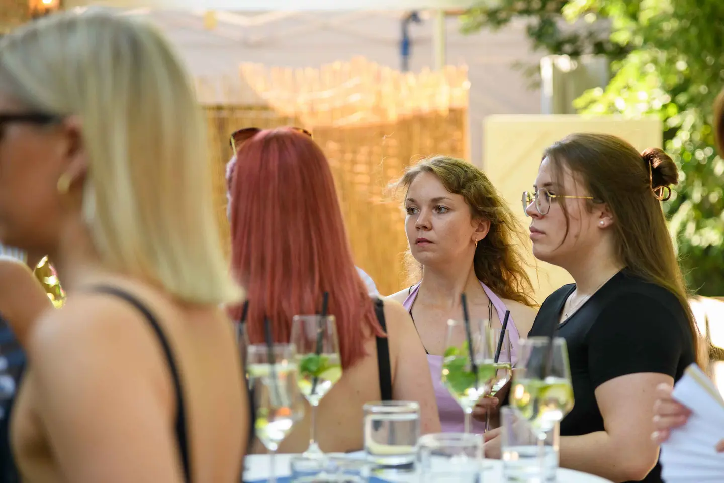 Junge Kolleginnen hören am Stehtisch aufmerksam zu, Weißweinspritzer davor – Sommerfest Eventfotografie Wien