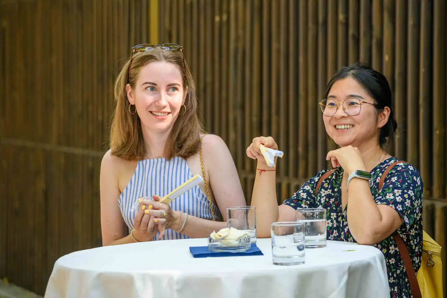 Zwei Kolleginnen am Stehtisch vor dunklem Bambuszaun, eine mit Fächer – Sommerfest Eventfotografie Wien