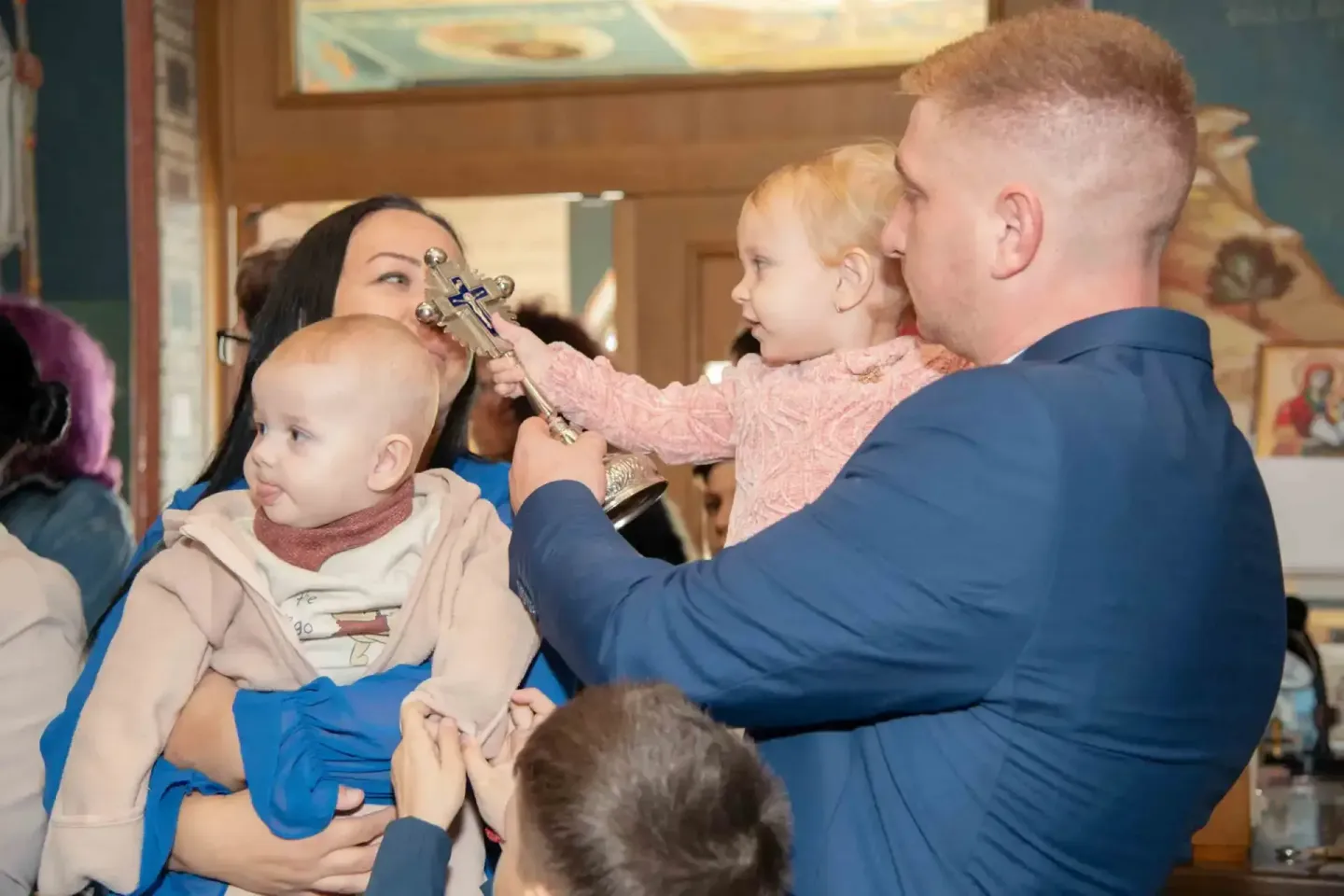 Eventfotograf Wien dokumentiert Taufe: Mann hält Kleinkind, das ein verziertes Kreuz betrachtet, während Frau daneben steht