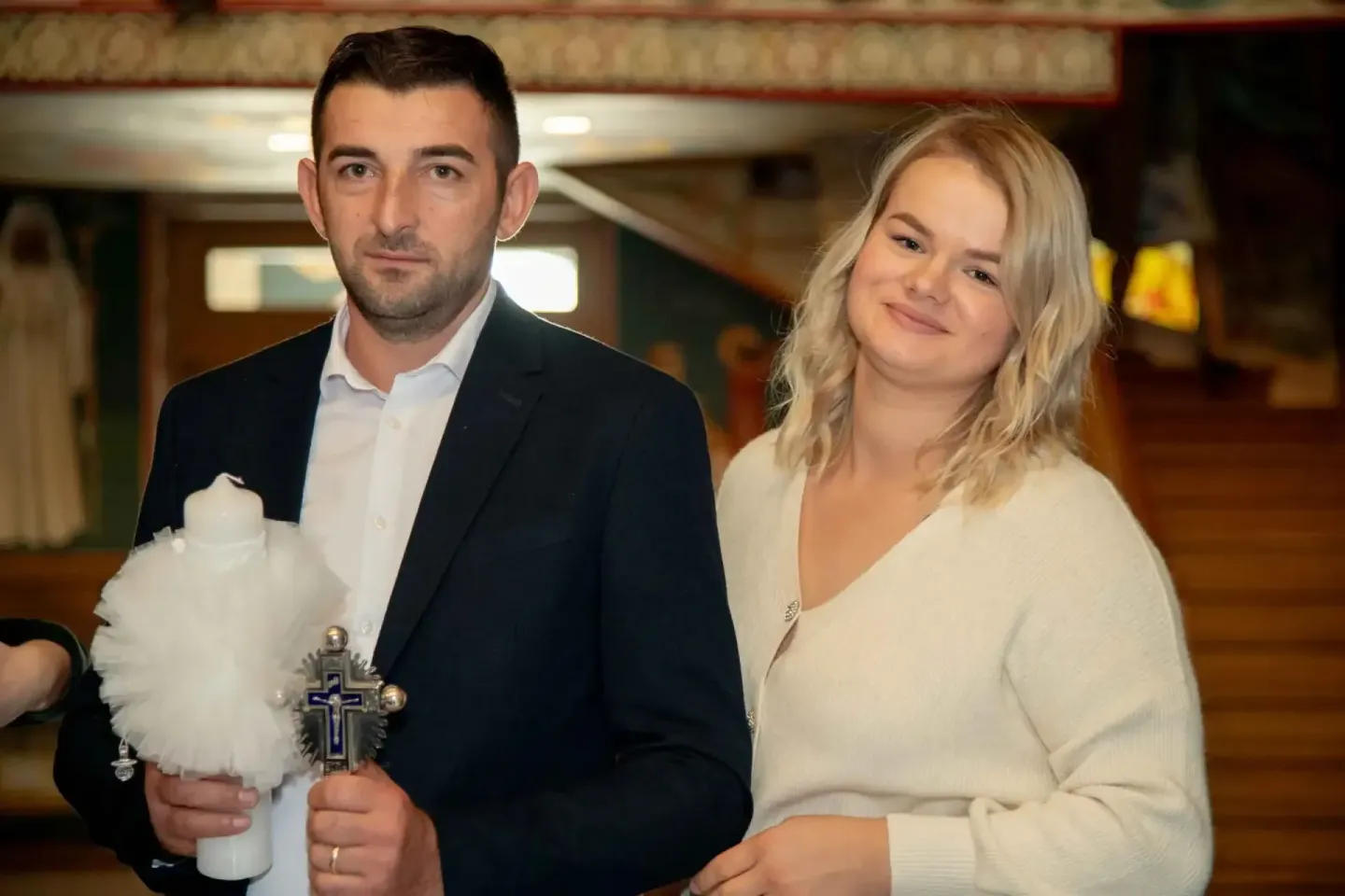 Eventfotograf Wien dokumentiert Tauffeier: Mann in dunklem Anzug und Frau in Creme-Cardigan halten Taufkerze