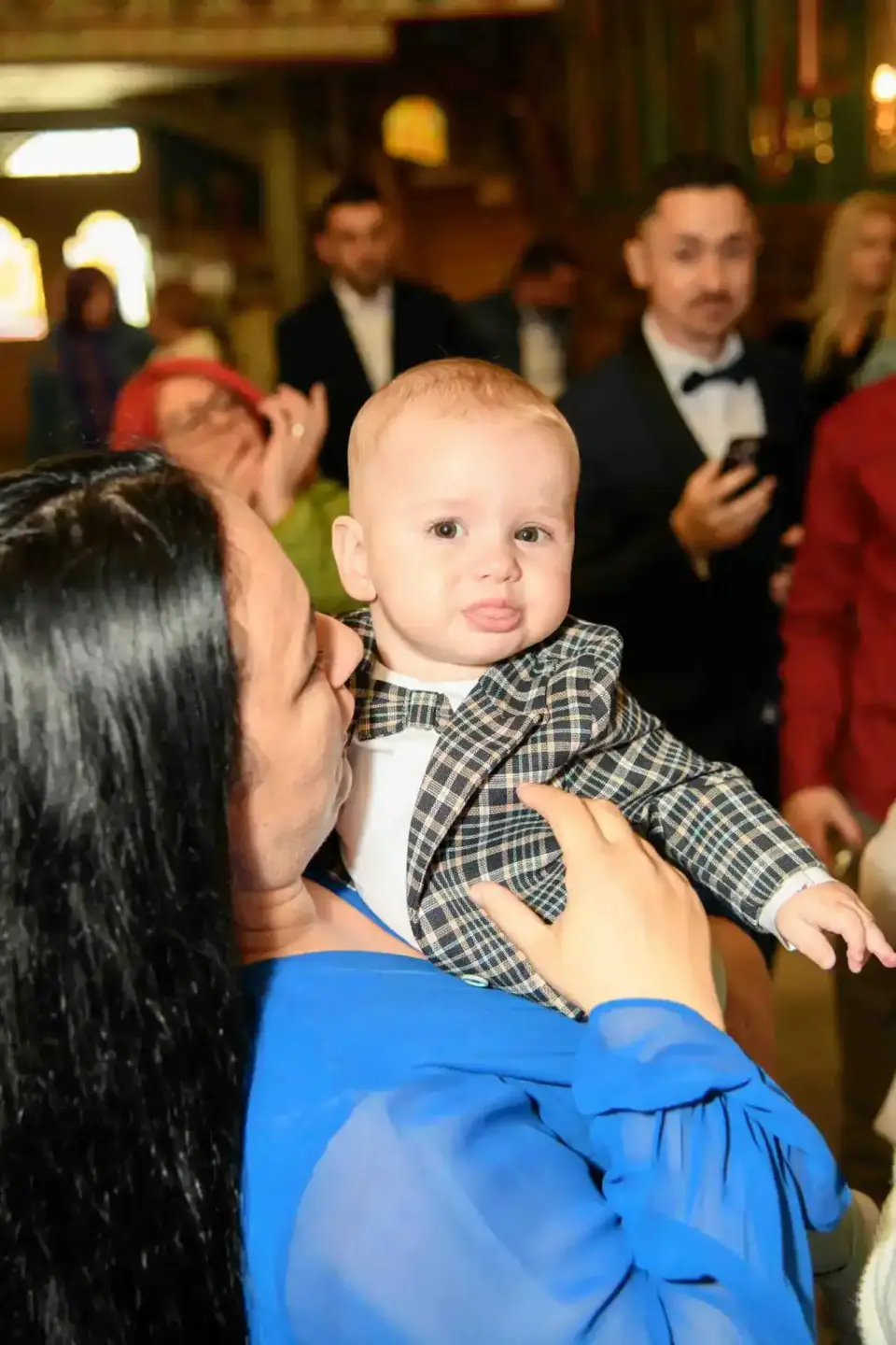 Eventfotograf Wien dokumentiert Taufe: Frau in blauem Kleid hält elegant gekleidetes Baby in kariertem Anzug, im Hintergrund 